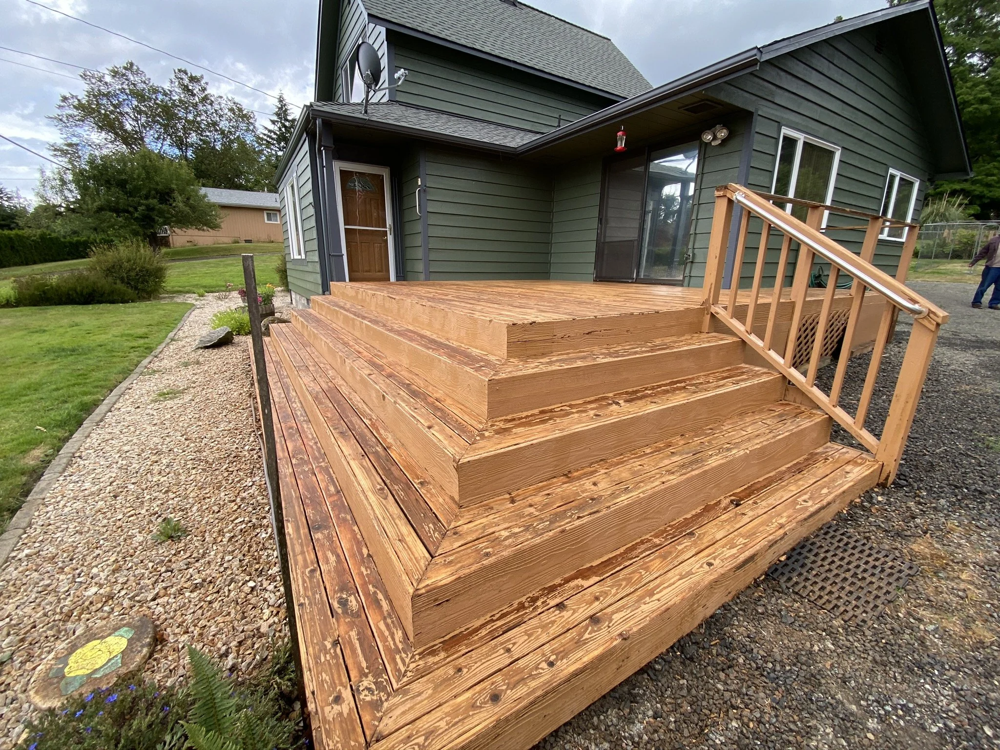 Newly built wooden deck with stairs and railing attached to the back of a green house, surrounded by a gravel walkway and a grassy yard with trees in the background.