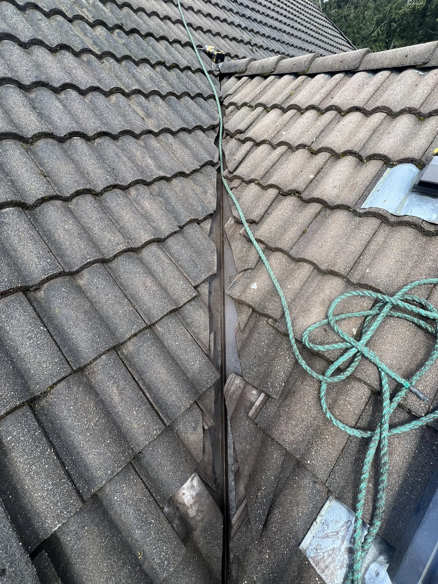 View of a roof with gray tiles. A black metal strip runs along the ridge, and there are ropes and a drill on the roof, indicating ongoing repairs or maintenance.