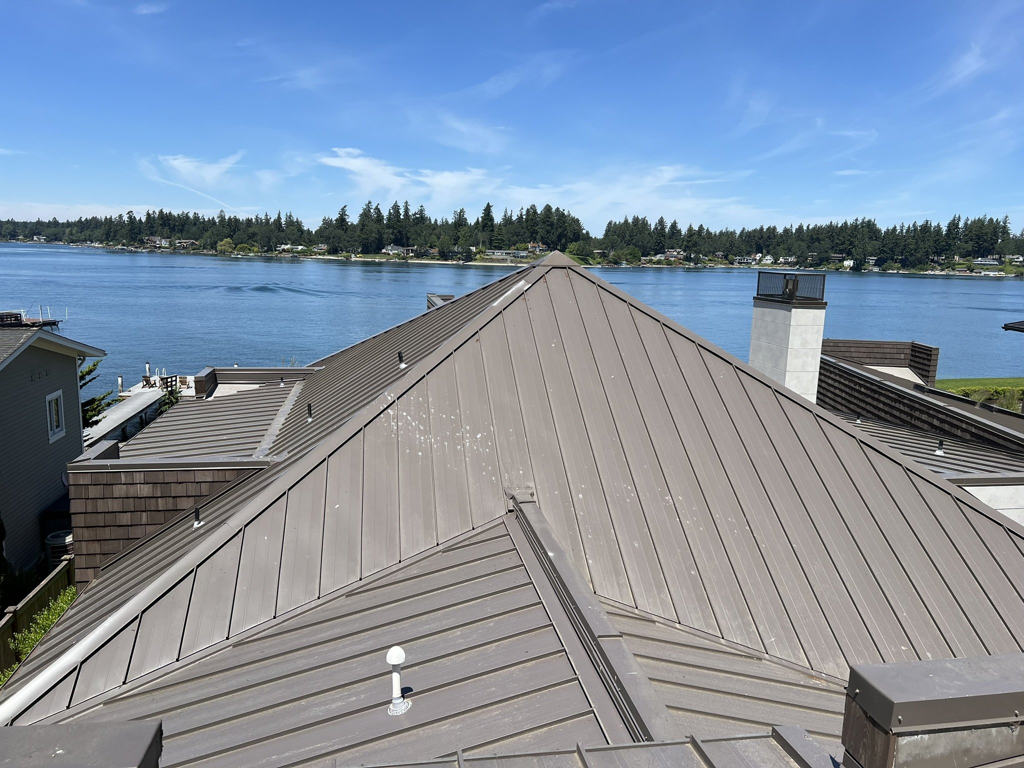 View over the rooftops of houses with a lake and a forested shoreline in the background under a blue sky with some clouds.