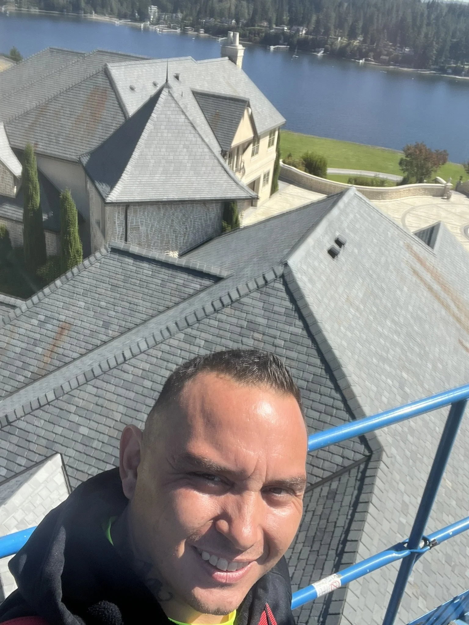 A man taking a selfie from a high vantage point showing the rooftops of houses below, a lake, and a green landscape in the background.