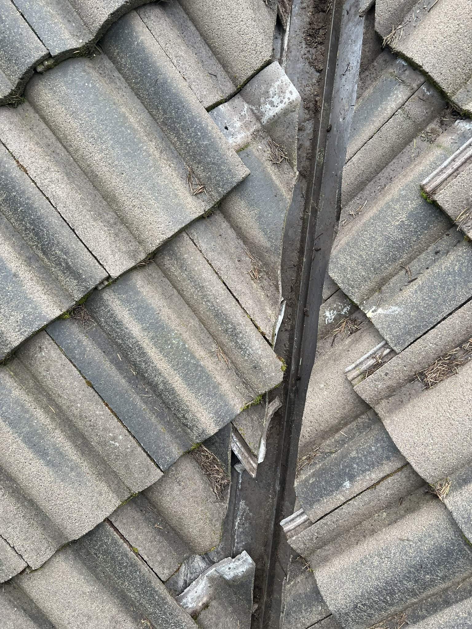 Close-up of a section of a tiled roof showing damaged tiles and a metal strip running down the center, with some moss and debris on the tiles.