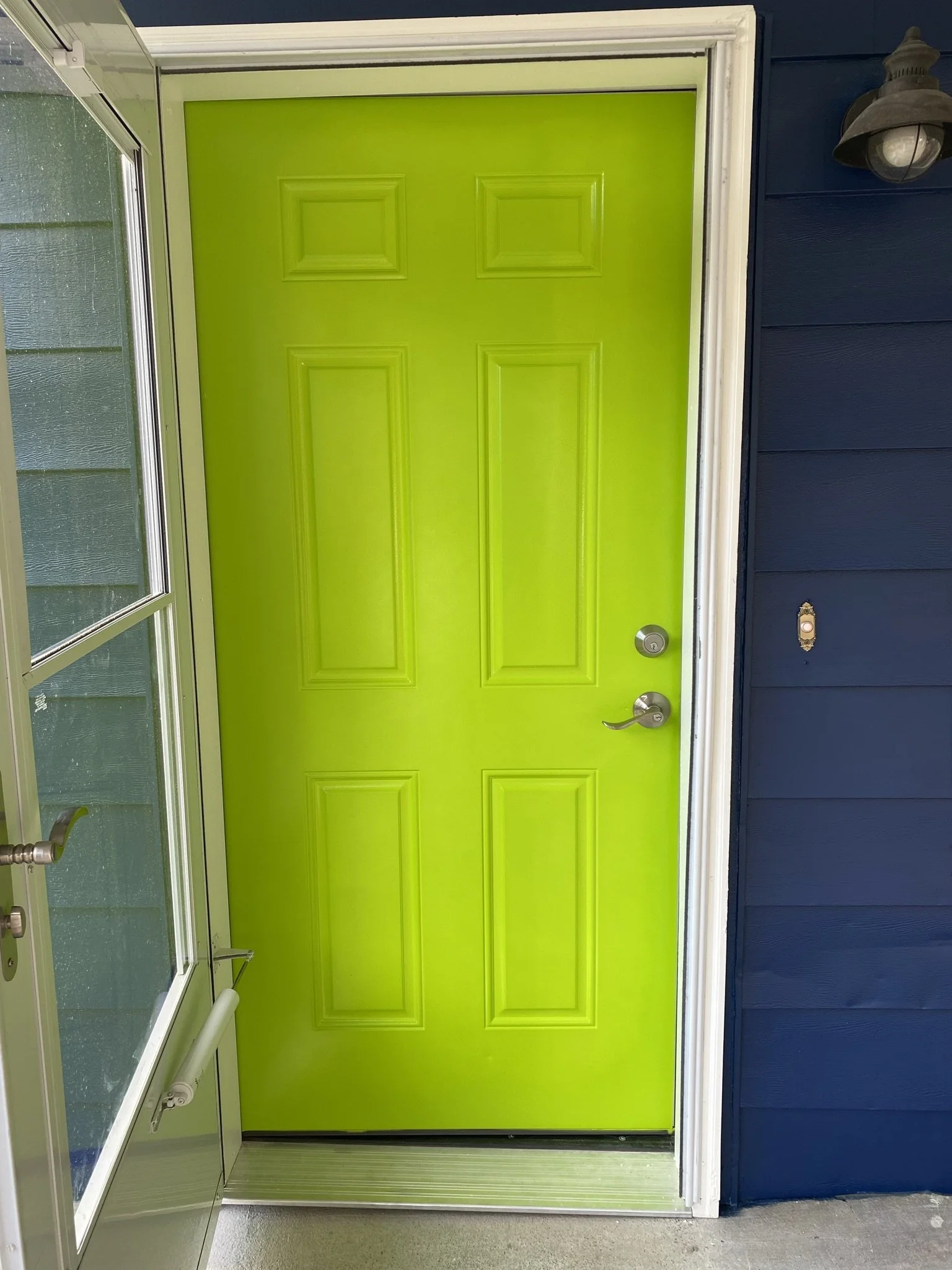 Bright lime green front door with a silver handle and lock, surrounded by white trim, next to blue house siding and a wall-mounted outdoor light fixture.