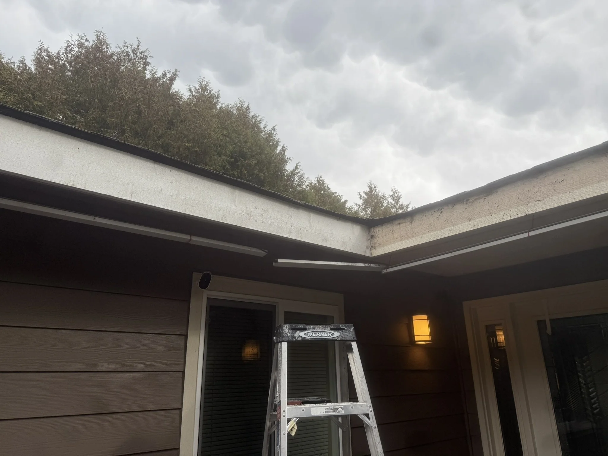 View of a house exterior with an open section of the roof showing exposed wood and a ladder nearby, under cloudy skies.