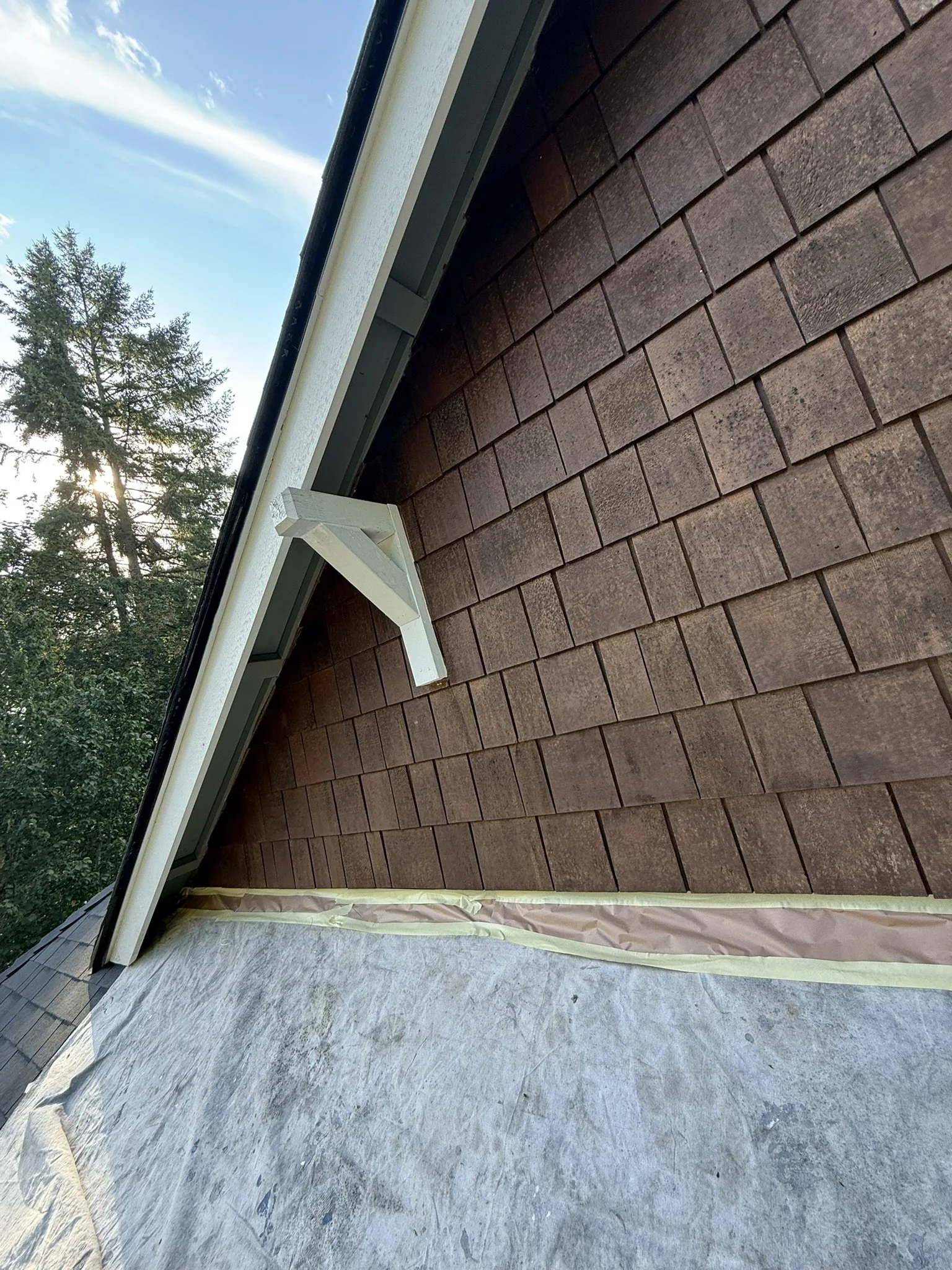 Close-up of a house's roof showing brown shingles, a white gutter, and an angled white support bracket, with a background of a clear blue sky and distant trees.