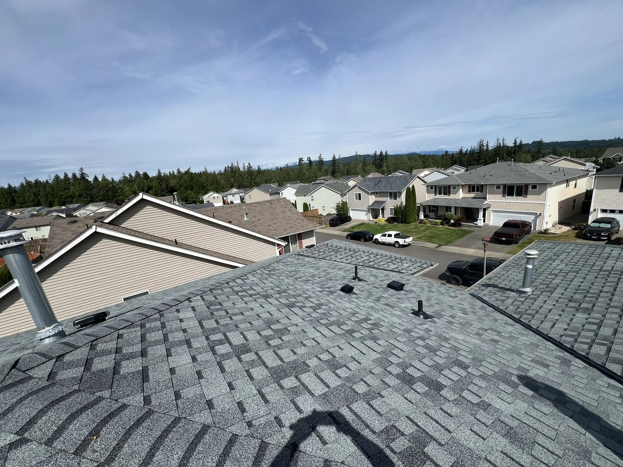 A suburban neighborhood with multiple two-story houses, a street with parked cars, and a backdrop of trees and distant mountains. The foreground shows rooftops with shingles, vents, and chimney pipes.