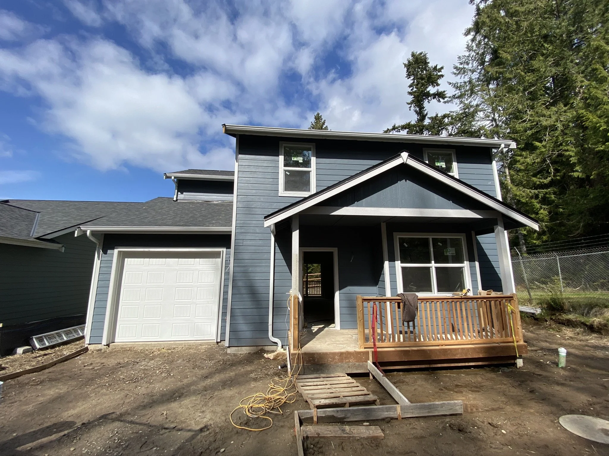 A two-story blue house with a front porch and a small staircase leading up to it. The house is surrounded by a dirt yard with some construction materials nearby. Trees are visible in the background, and the sky is partly cloudy.