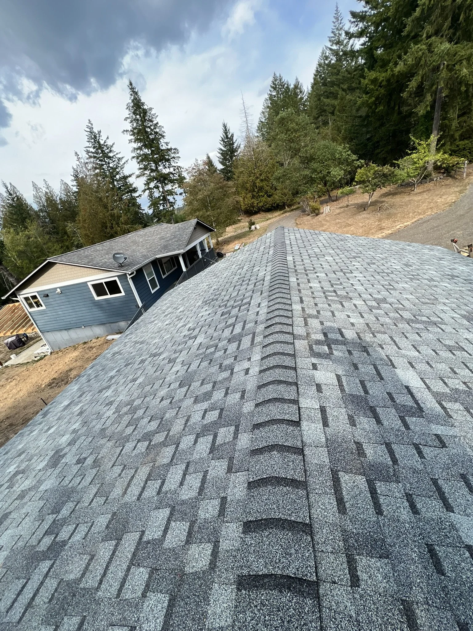 A roof with gray shingles, viewed from above, with a house and trees in the background under a partly cloudy sky.
