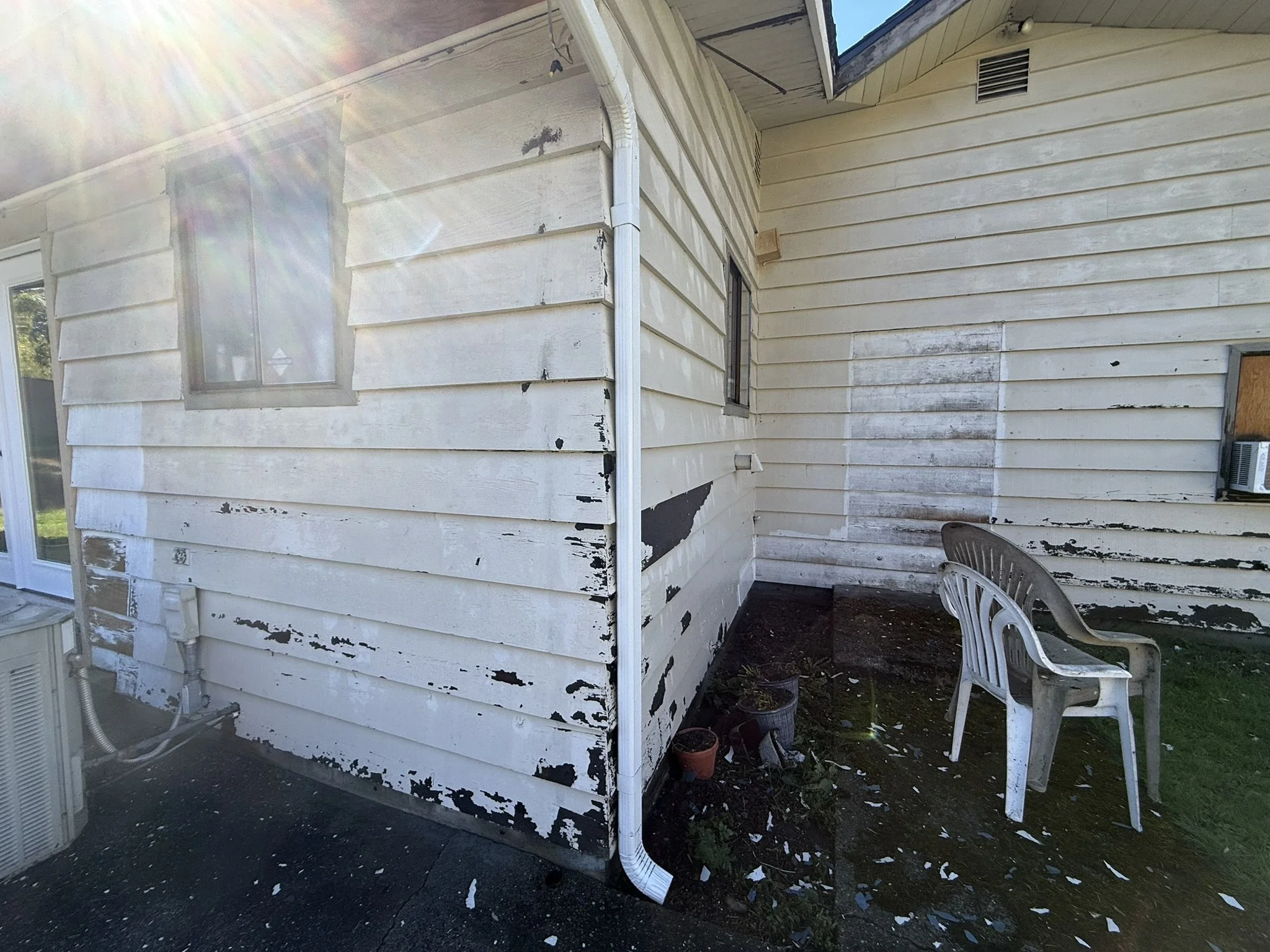 Exterior of a house with peeling white paint on the siding, a plastic chair, and a window air conditioning unit.
