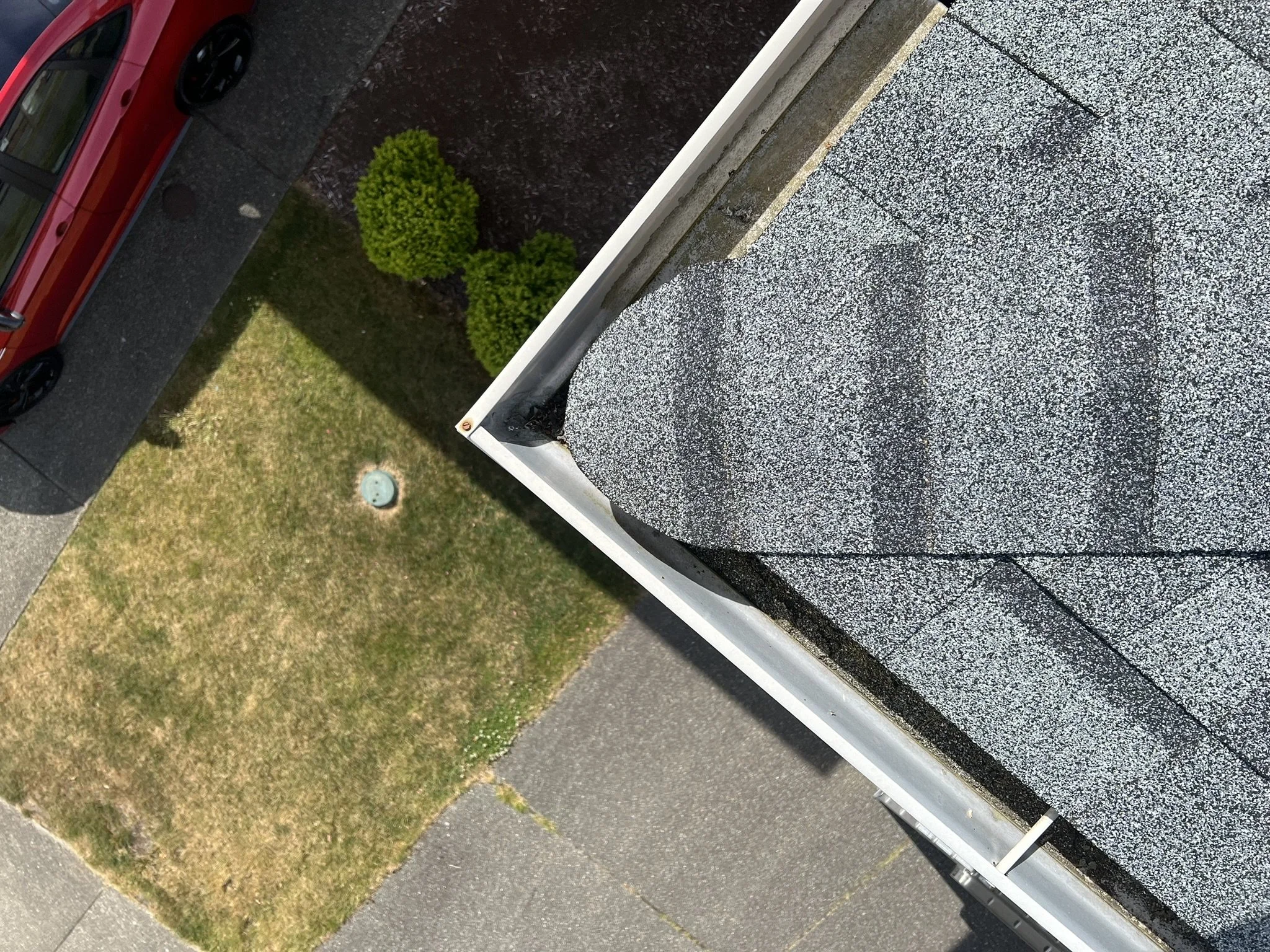 View from above shows a gray asphalt roof with black cracks, a white gutter, a patch of trimmed green bushes, a small grassy area with some patchy yellow grass, and part of a sidewalk and street with parked cars.