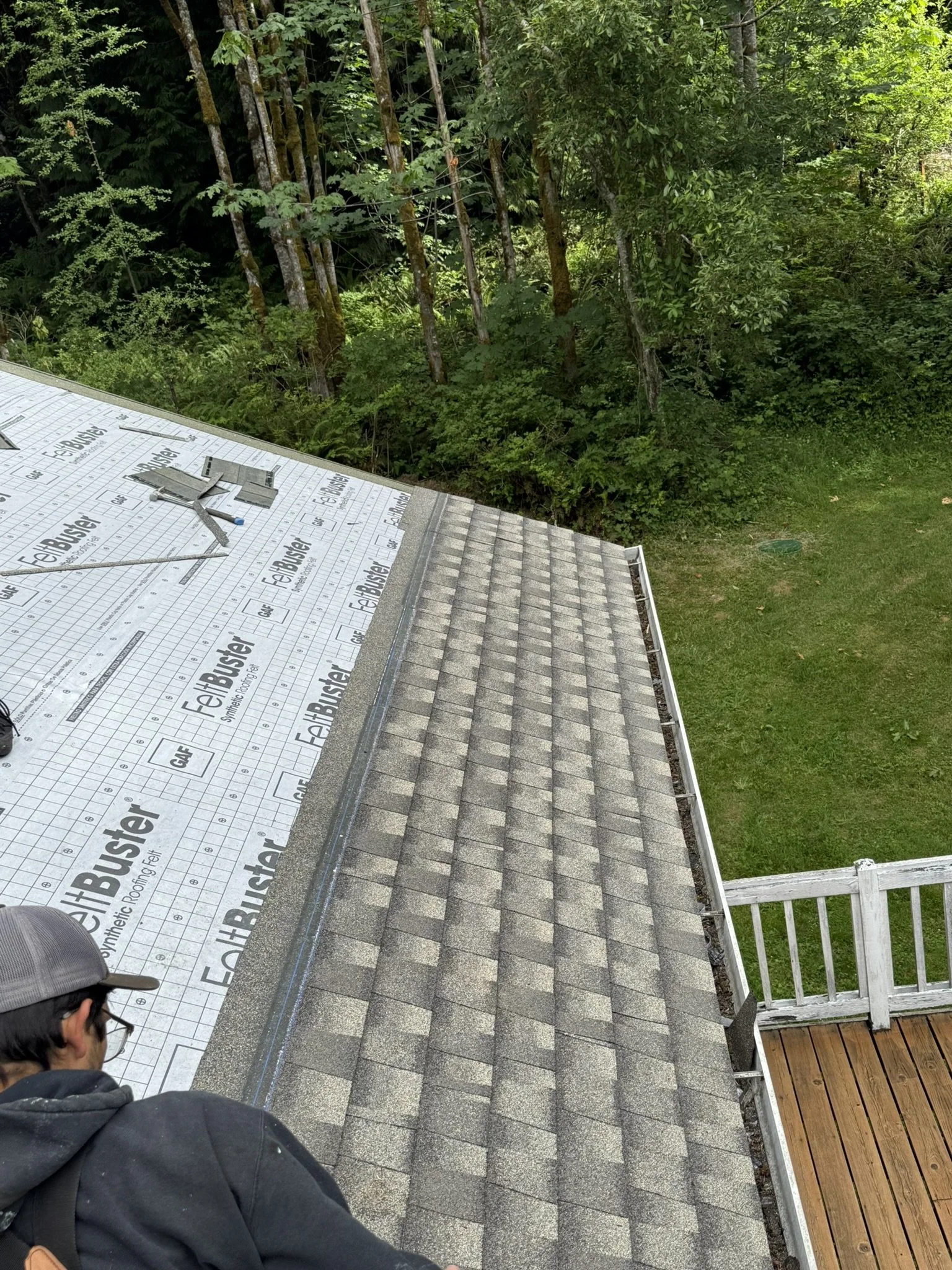 View of a roof under construction with asphalt shingles, partially covered with synthetic roof underlayment, and a worker in the lower left corner. There is a deck with a railing at the right side, and green trees and grass in the background.