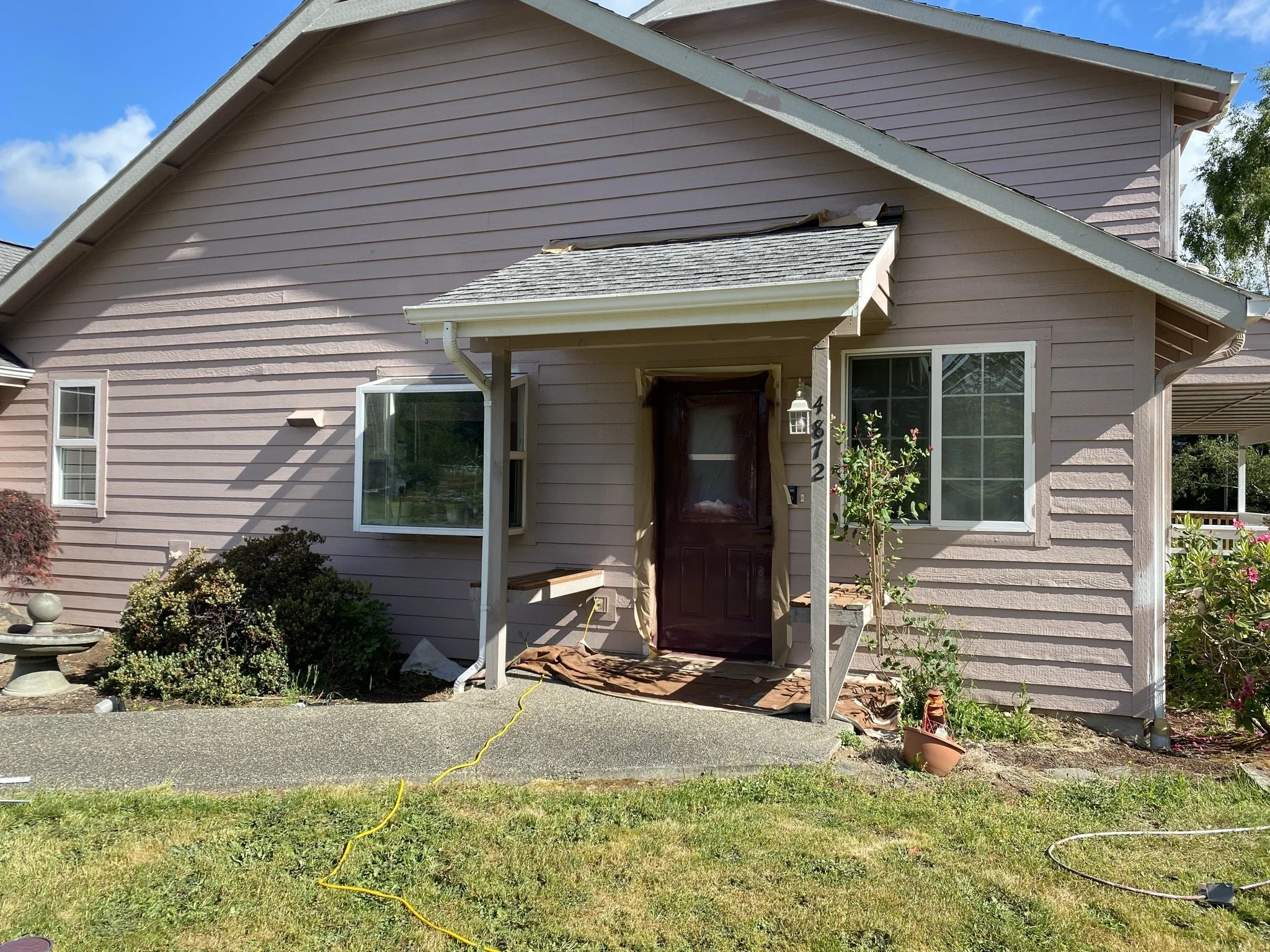 A house with pink horizontal siding, a small front porch, and a brown front door. There is a window with a bay window design on the left and another window on the right. The front yard has bushes, a small bird bath, potted plants, and a garden hose o