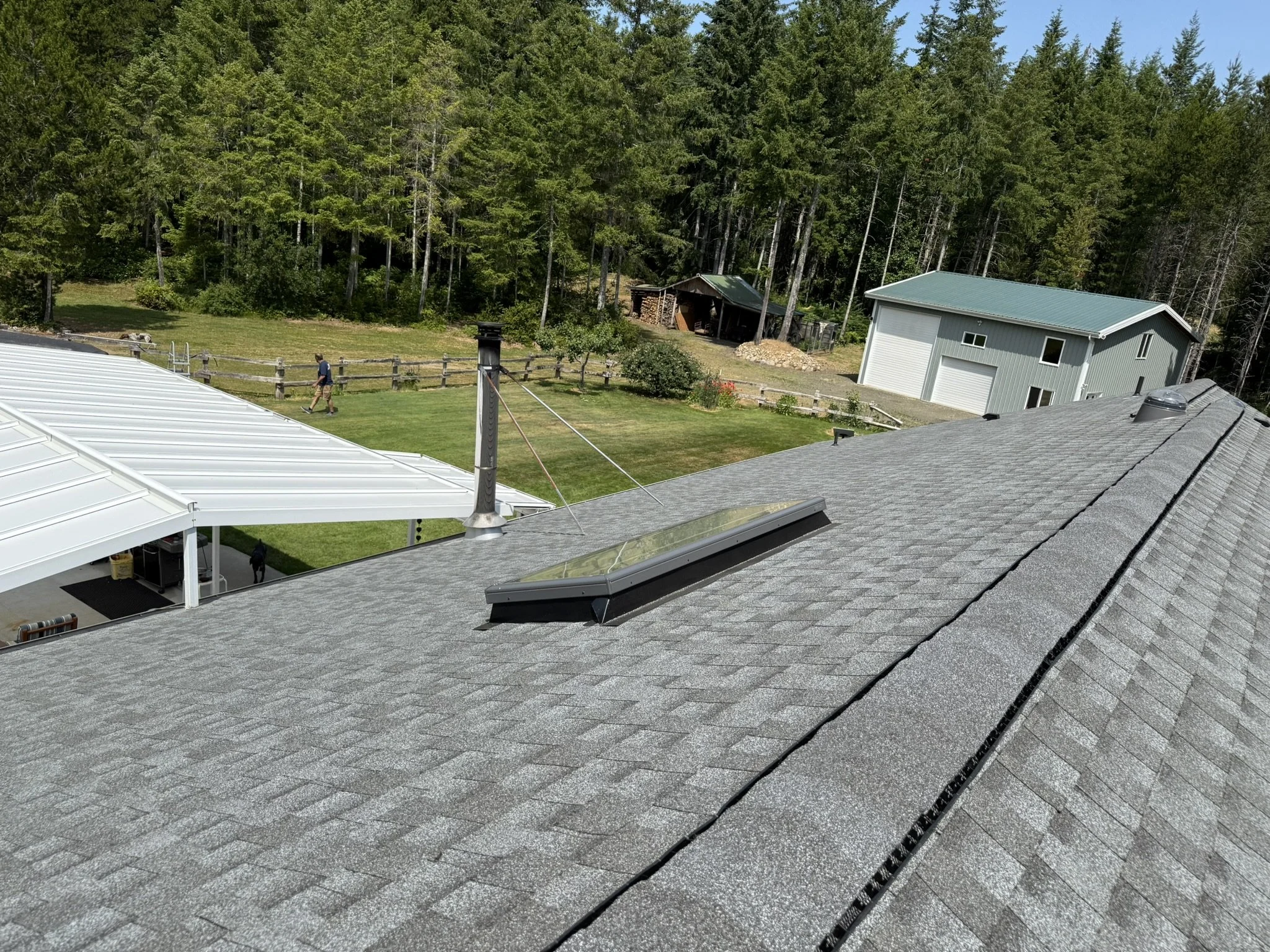 A view from a roof showing a gray shingled roof with a skylight, a chimney with a ventilation cap, and a backyard with green grass, a person walking, and various trees and structures in the distance.