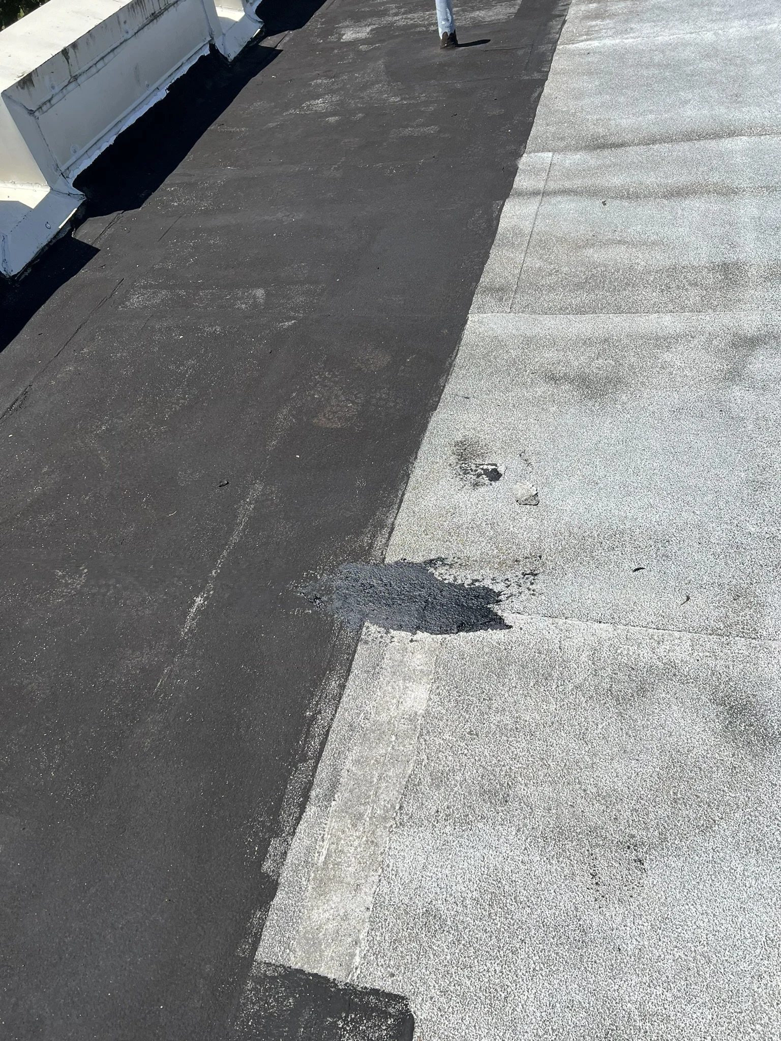 Section of a roof with black and gray asphalt roofing materials, showing a small patch of newer black material on the gray surface.