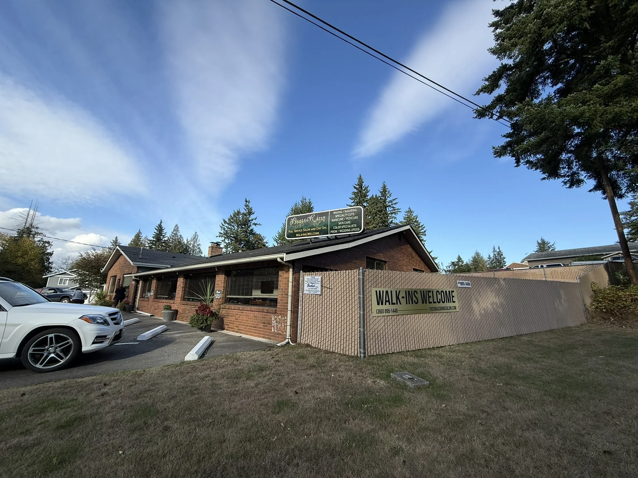 A brick building with a parking lot in front, some cars, and a sign that reads 'WALK-INS WELCOME' with a phone number. There are trees and a blue sky with scattered clouds.