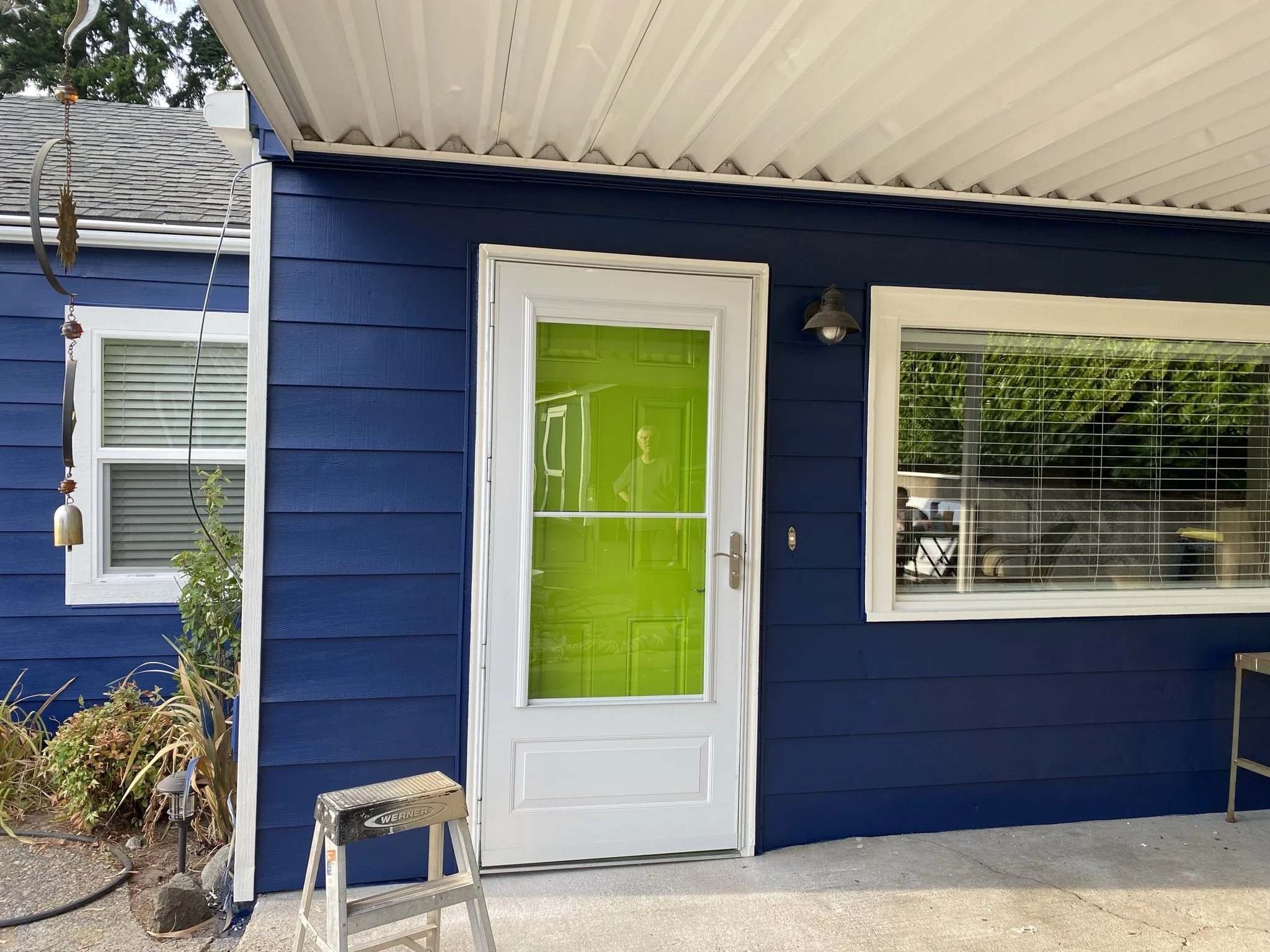 Front of a house painted in dark blue with white trim, featuring a white door with a large green window and a neighboring window with blinds, outdoor plants, and a small ladder on the concrete porch.