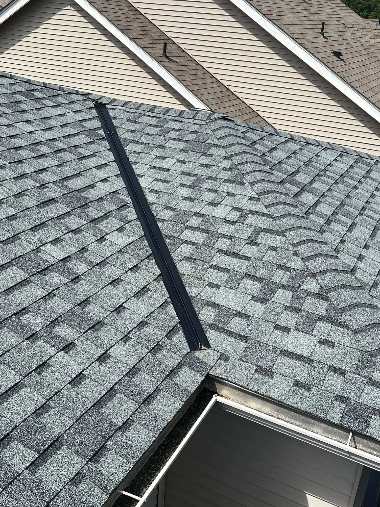 Close-up view of a house roof with gray asphalt shingles, black metal vent pipe, and a part of a beige house wall with siding.