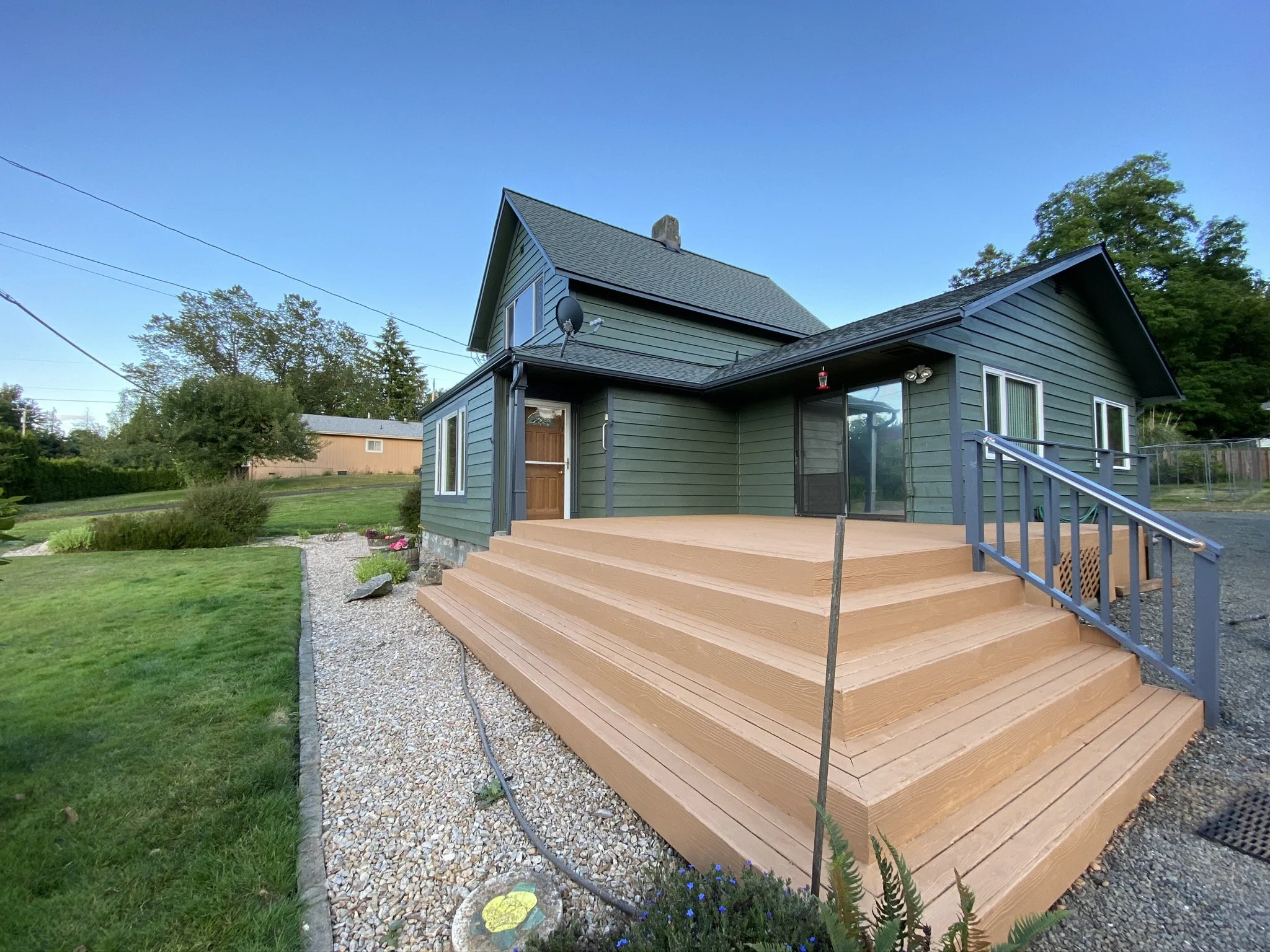 A house with dark green siding and a new wooden deck with stairs, located in a green yard with trees in the background.
