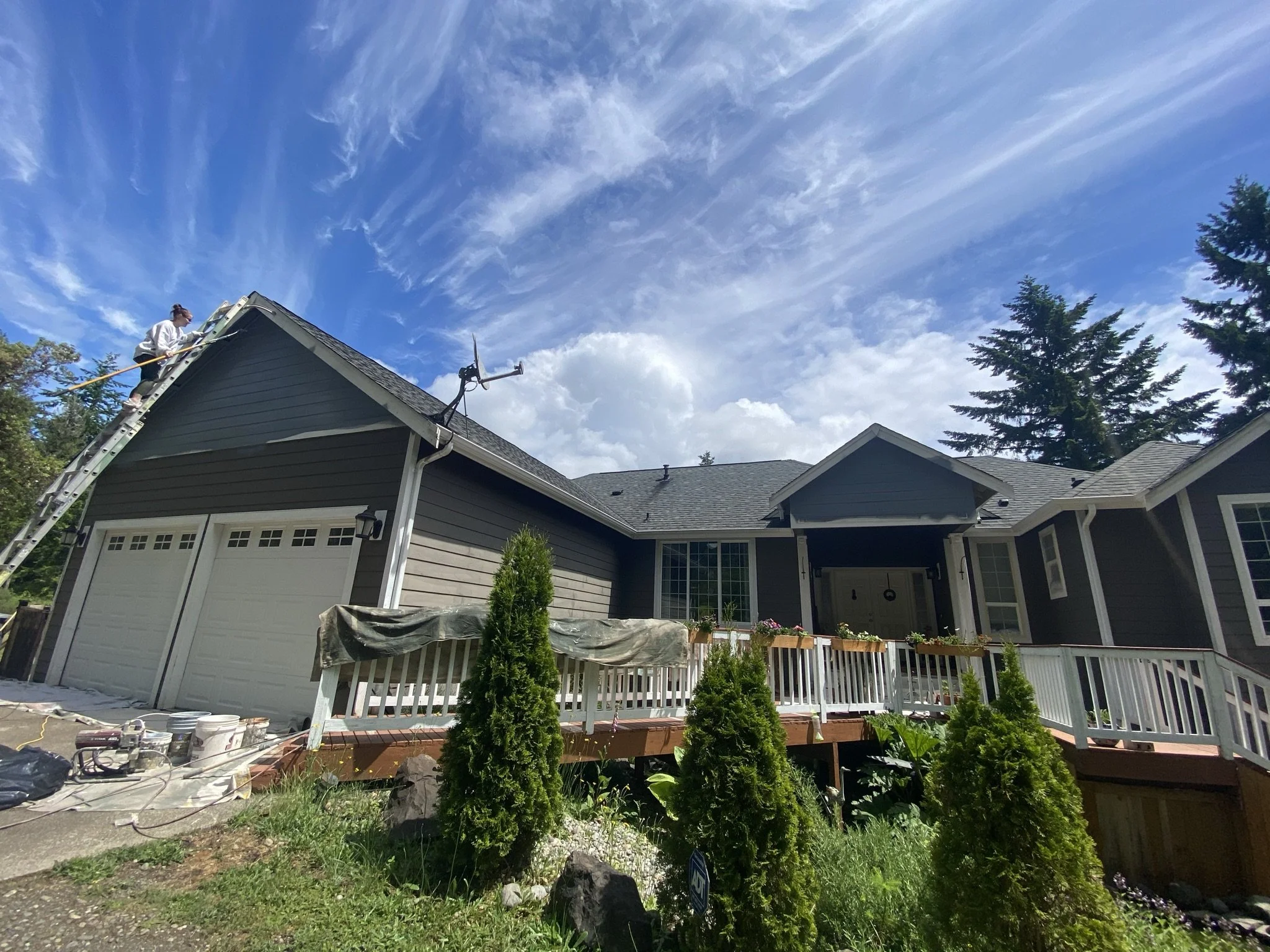 A house with dark gray siding and a white garage door is shown with a crew member on a ladder working on the roof. There is a deck with potted plants and a row of small evergreen shrubs in the front yard. The sky is blue with white clouds.