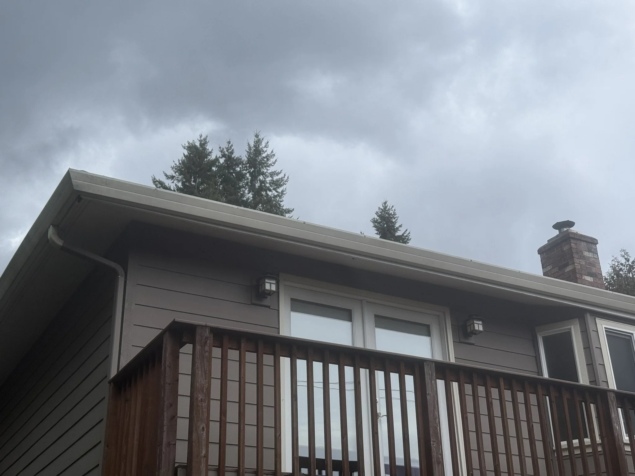 The image shows the upper part of a house with a wooden balcony, grey siding, and a chimney under a cloudy sky.