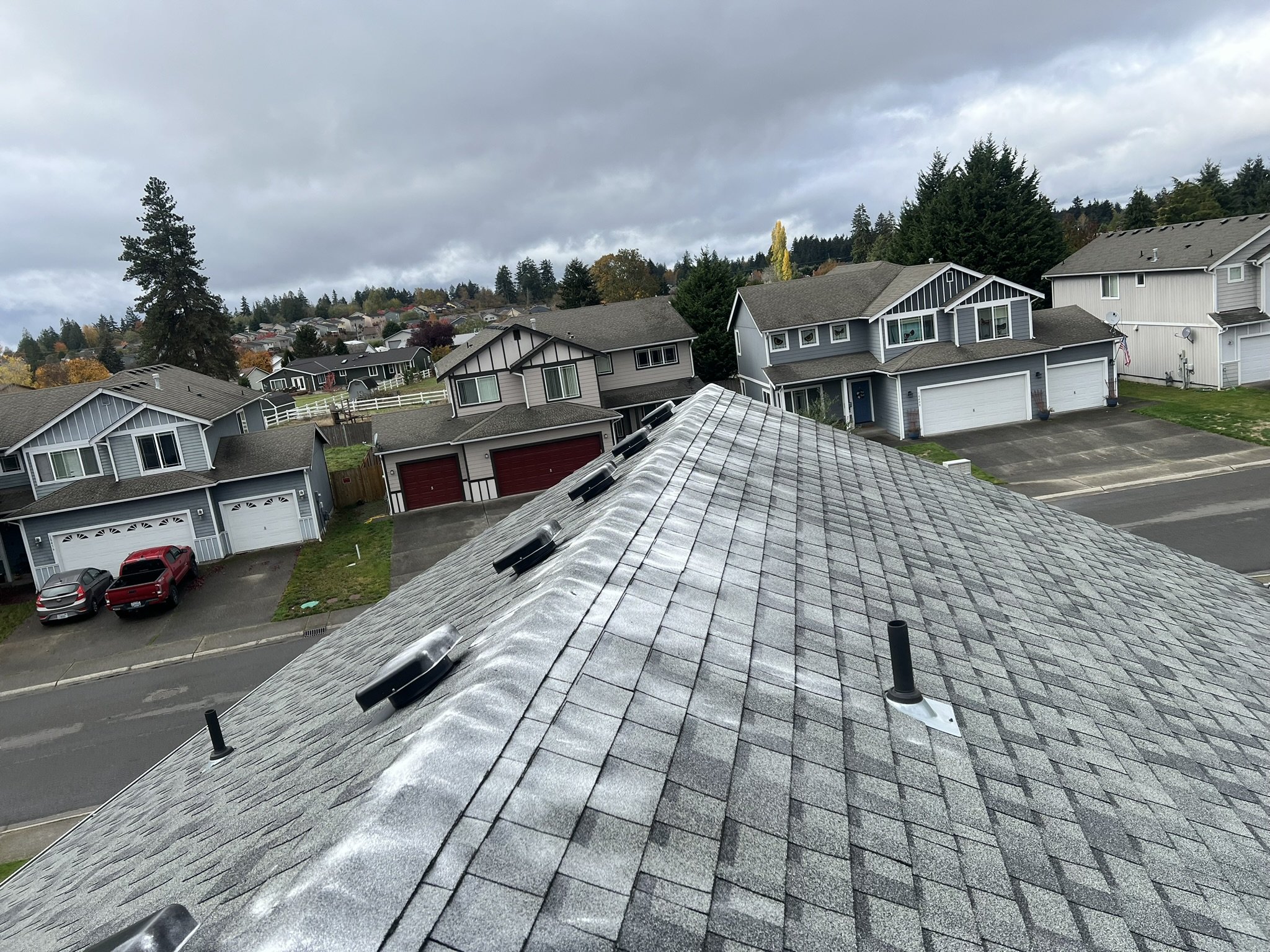 View from a roof showing neighboring houses, some with garages, on a cloudy day. Multiple vents and flashing are visible on the roof.