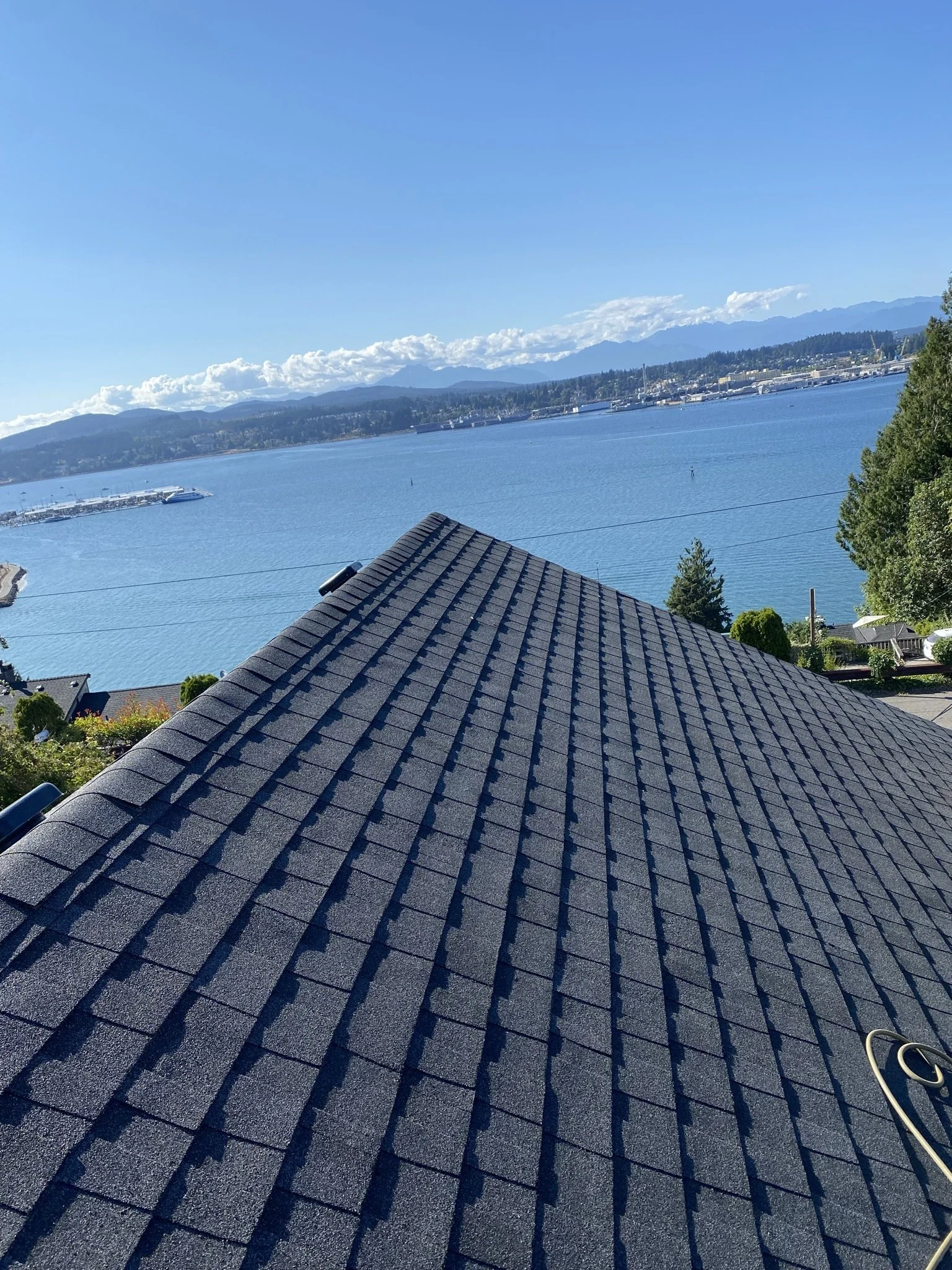 Looking over a house roof with dark gray shingles, overlooking a body of water with docks, hills, and mountains under a partly cloudy blue sky.