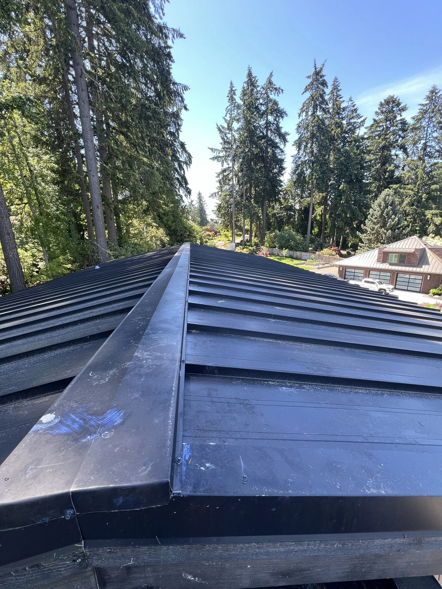 A metal roof with black panels extending towards trees and houses, with a central ridge running along the top.
