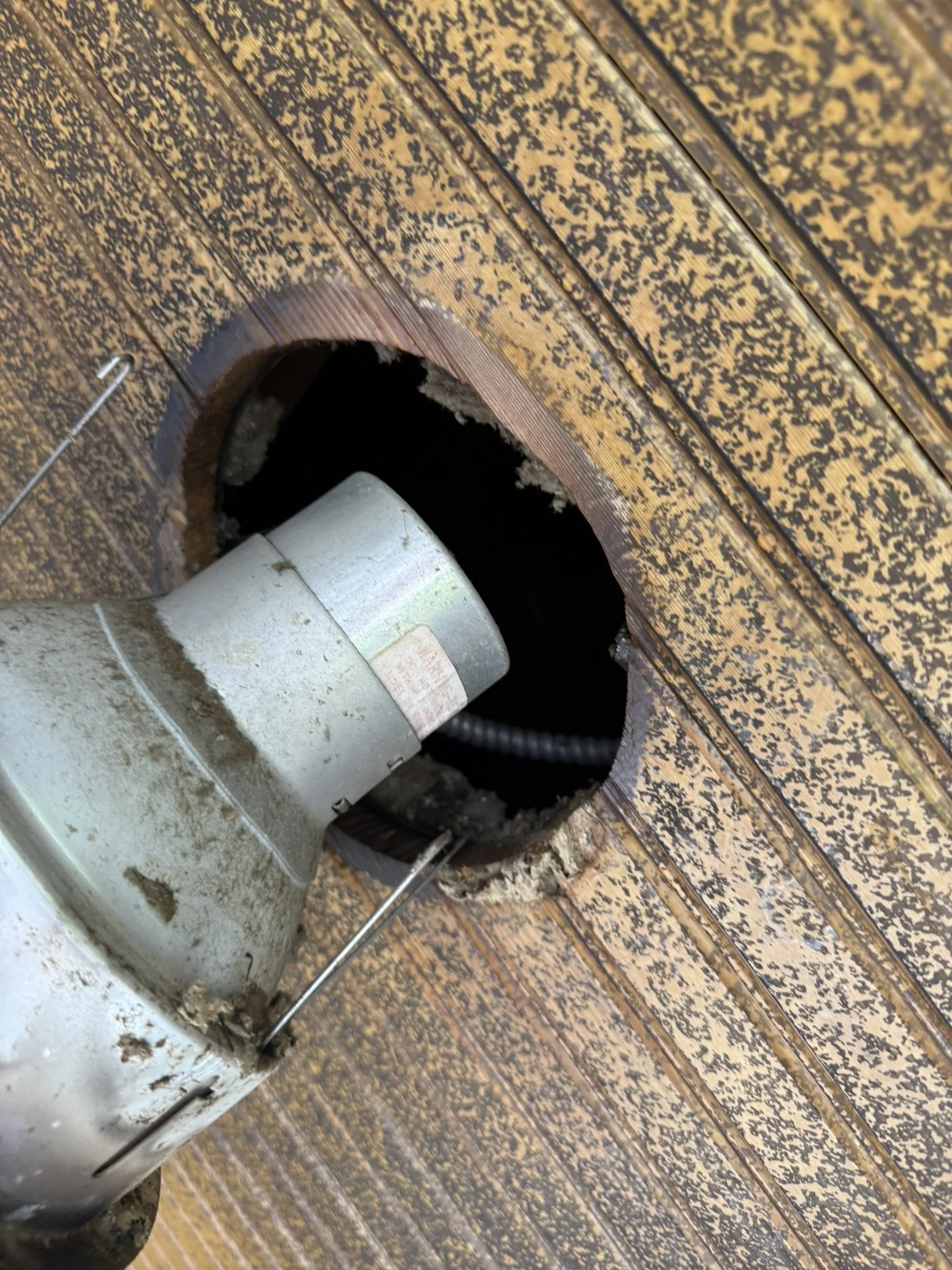 Close-up of a hole in a wooden ceiling or wall with a motor or fan unit partially visible inside the hole.