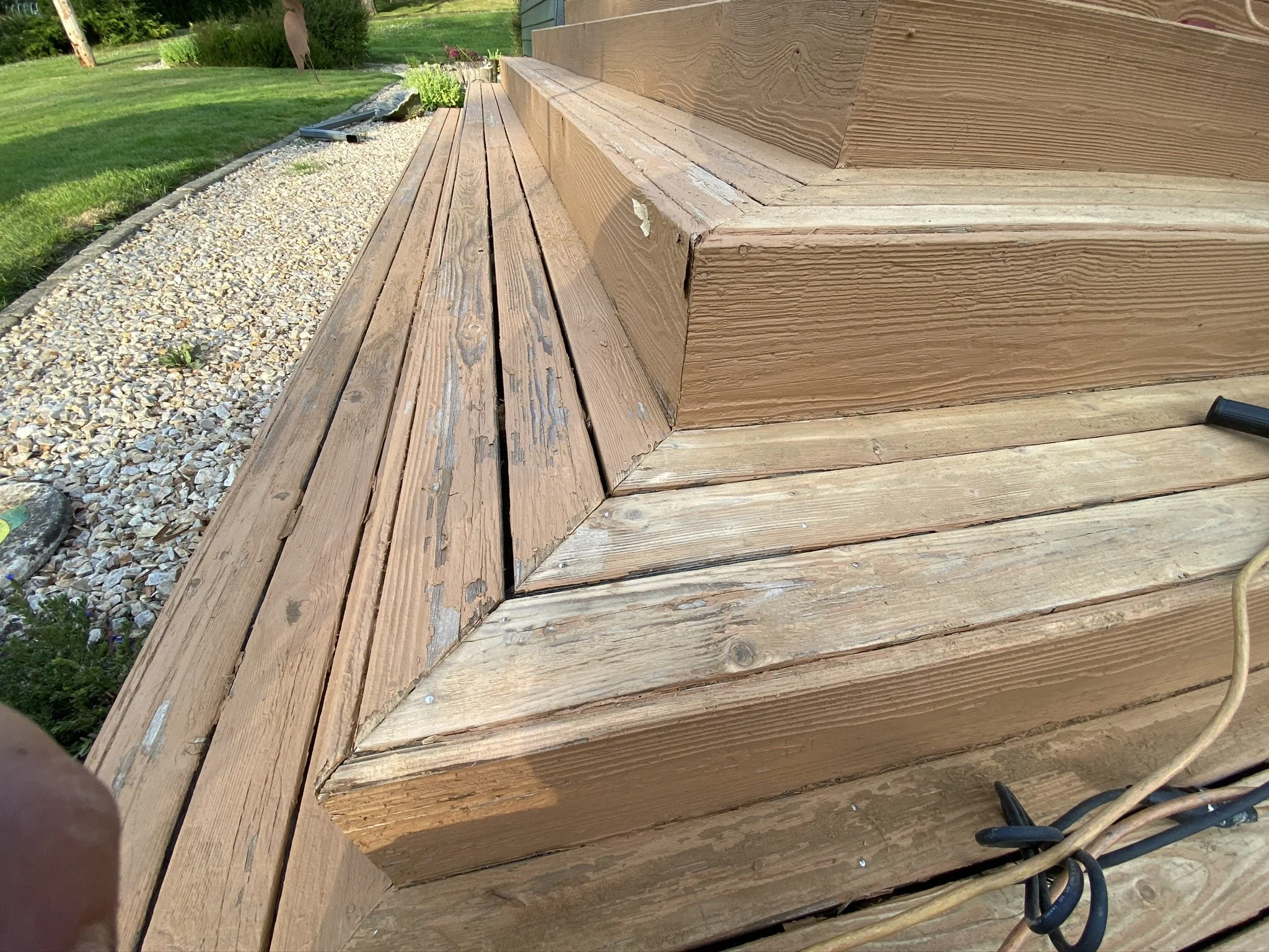 A close-up of a wooden deck and steps, with some weathered and peeling paint on the wood. The deck is next to a grassy yard with a gravel border and garden plants.