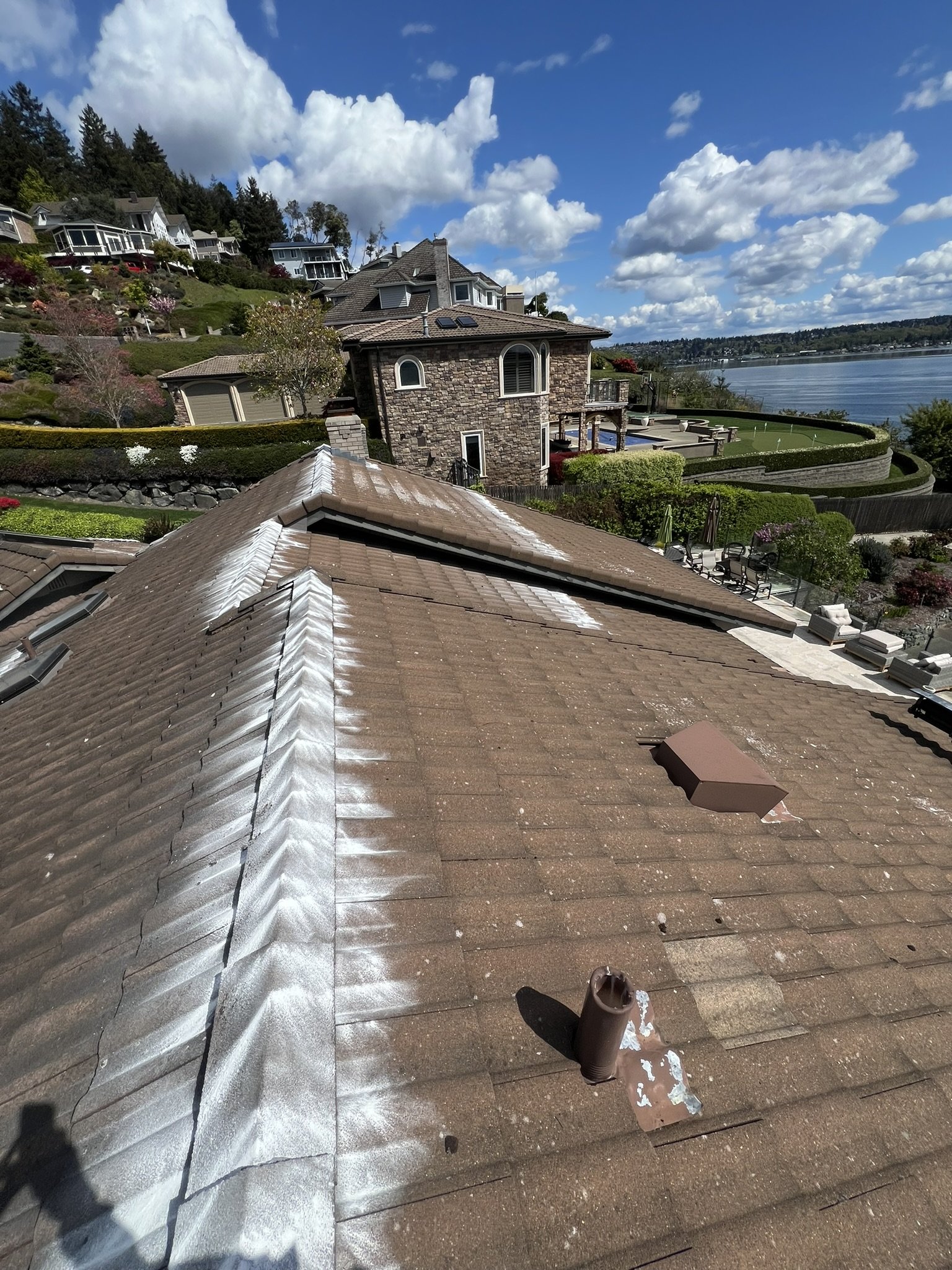 View of a residential rooftop with white sealant along the tagged seams, overlooking a neighborhood with large houses, a hillside, a lake, and a blue sky with white clouds.