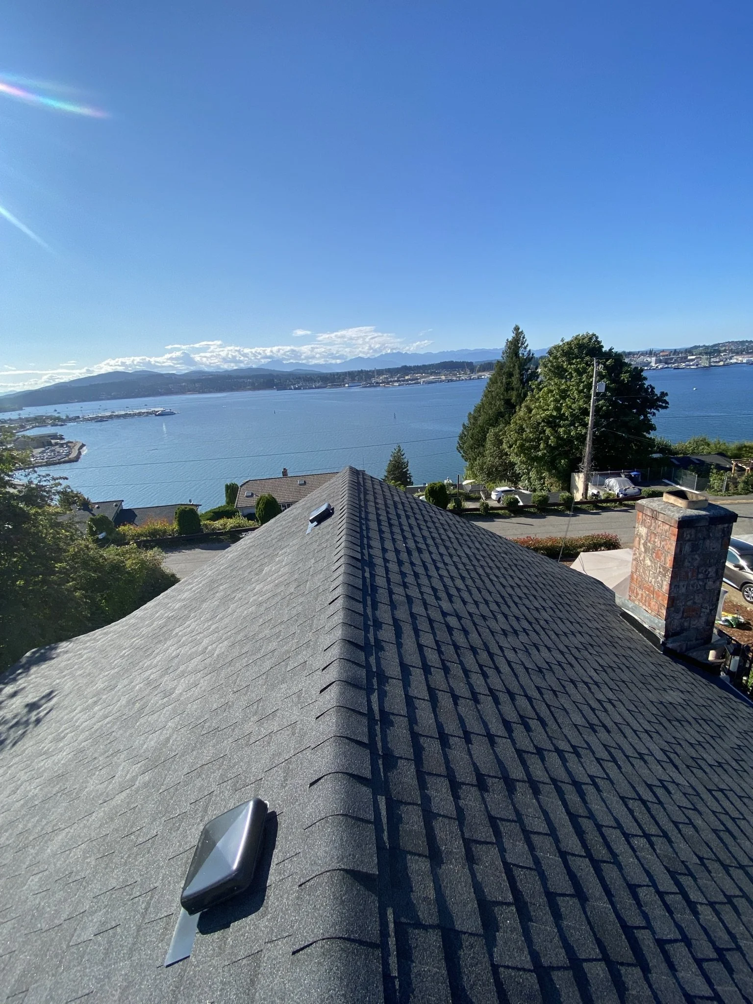 View from the roof of a house overlooking a body of water, with trees, cars, and houses nearby, under a clear blue sky.