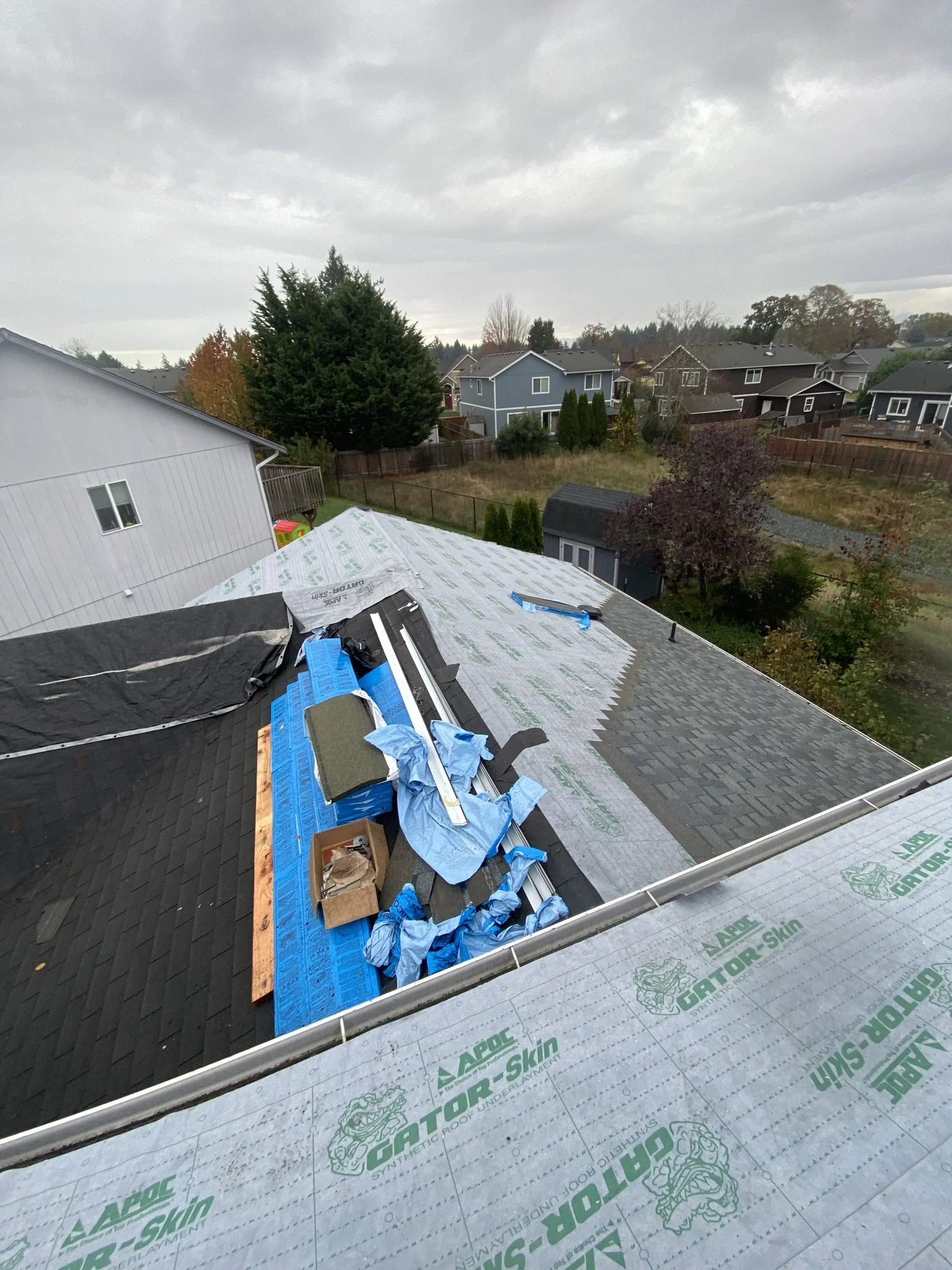 View of a house roof under construction, with roofing materials and tools, and a neighborhood with houses and trees in the background under cloudy sky.
