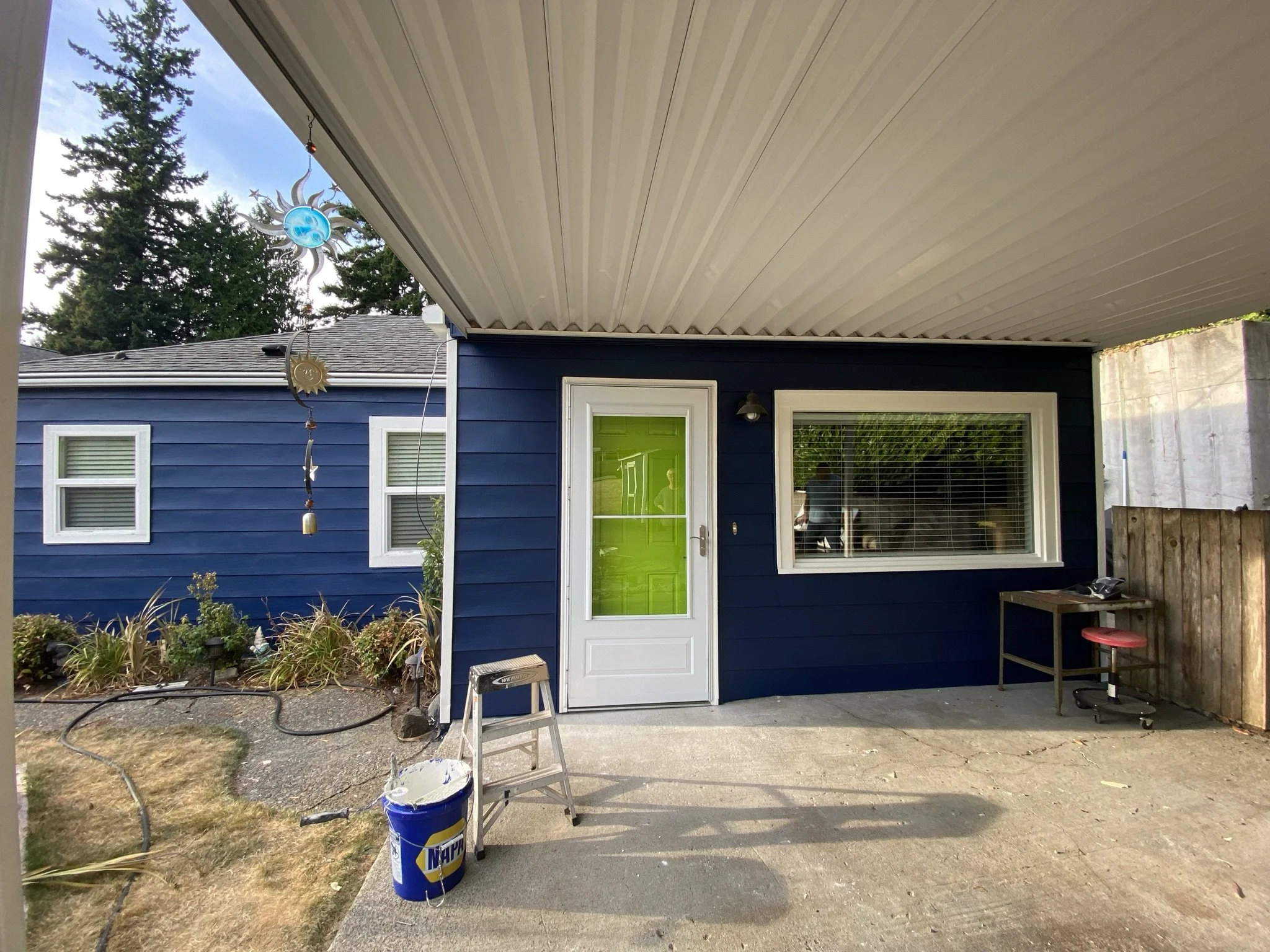 Back patio of a house with a blue exterior, white door with a neon green window, window with blinds, and a ceiling with a solar sun decor hanging. There is a ladder, a paint bucket, a small table, and a red stool on the concrete patio.