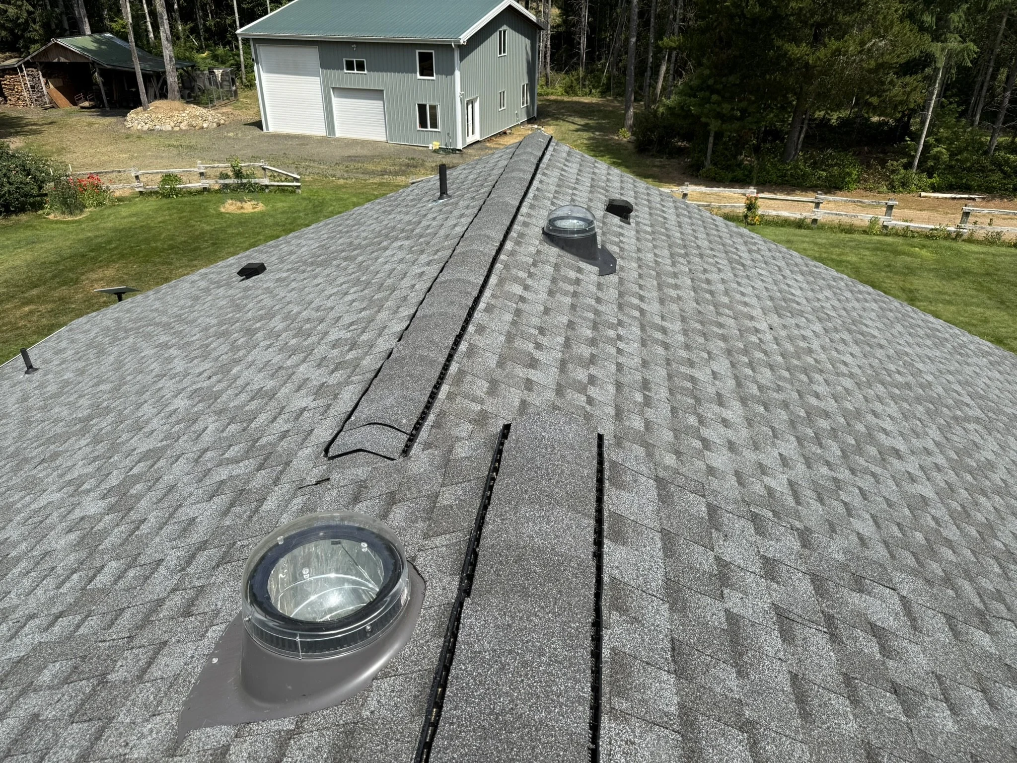 View of a residential roof with gray asphalt shingles, roofing vents, and a skylight, overlooking a backyard with a green lawn, flower beds, and a gray house.
