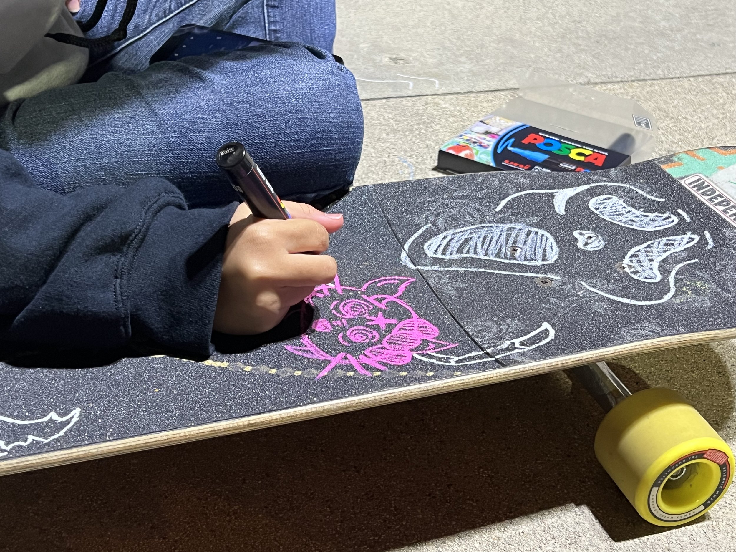 Person in a black hoodie drawing on a skateboard with colored chalk. The skateboard has colorful drawings, including a pink cartoon pig. A box of Posca paint markers is on the ground next to the skateboard.