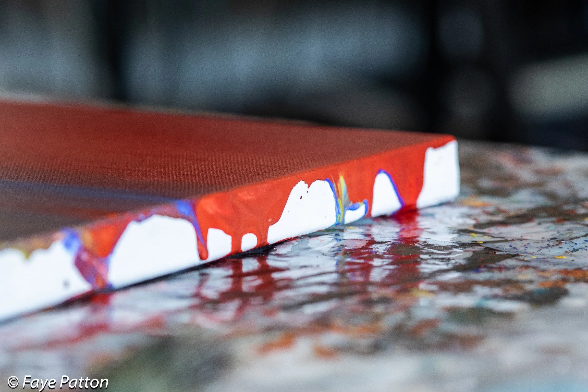 Close-up of a canvas with red paint and white areas that have colorful abstract designs on its edge, resting on a textured, multicolored surface.