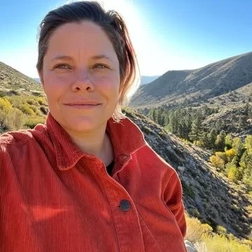 Person taking a selfie outdoors in a mountainous area with clear blue sky, wearing a red jacket.