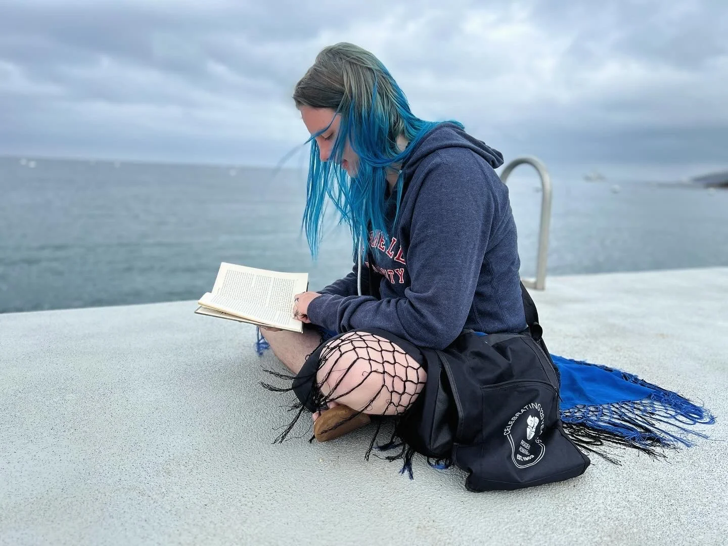 Young woman with blue hair reading a book while sitting on a concrete pier near the water, wearing fishnet tights and a hoodie, with a black bag beside her on a blue blanket.