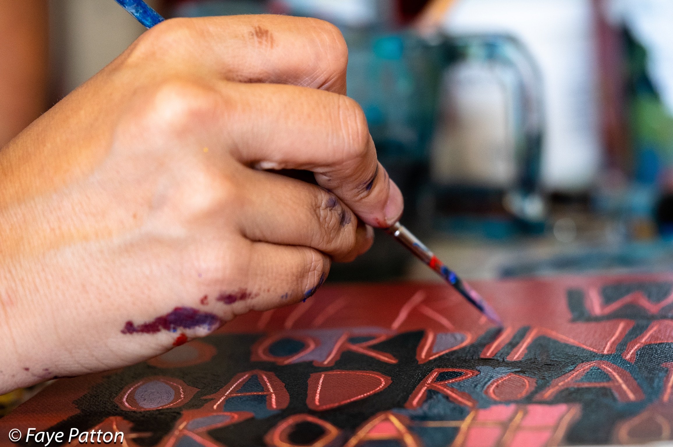 Close-up of an artist's hand holding a small paintbrush, painting colorful letters and shapes on a canvas.