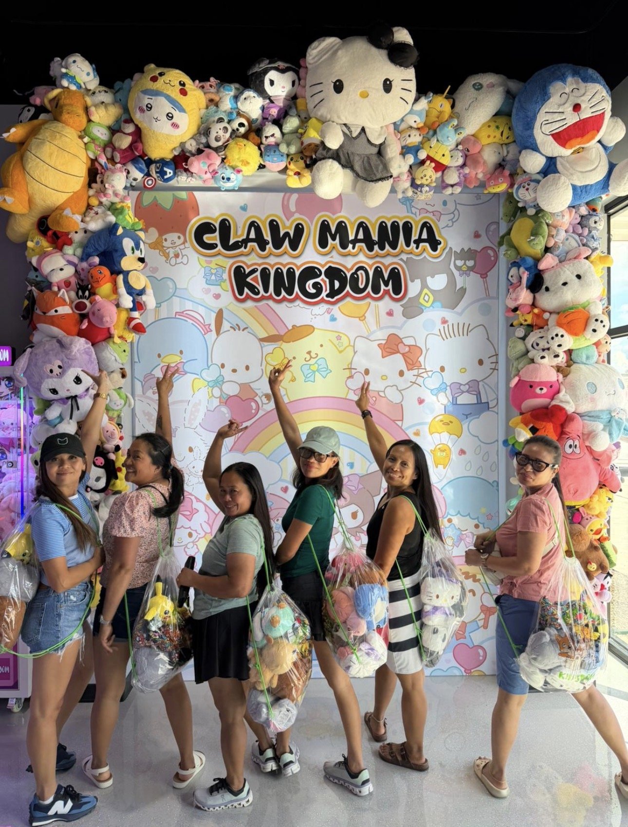 Group of five women posing in front of a colorful display of plush toys at Claw Mania Kingdom, cheering and smiling.