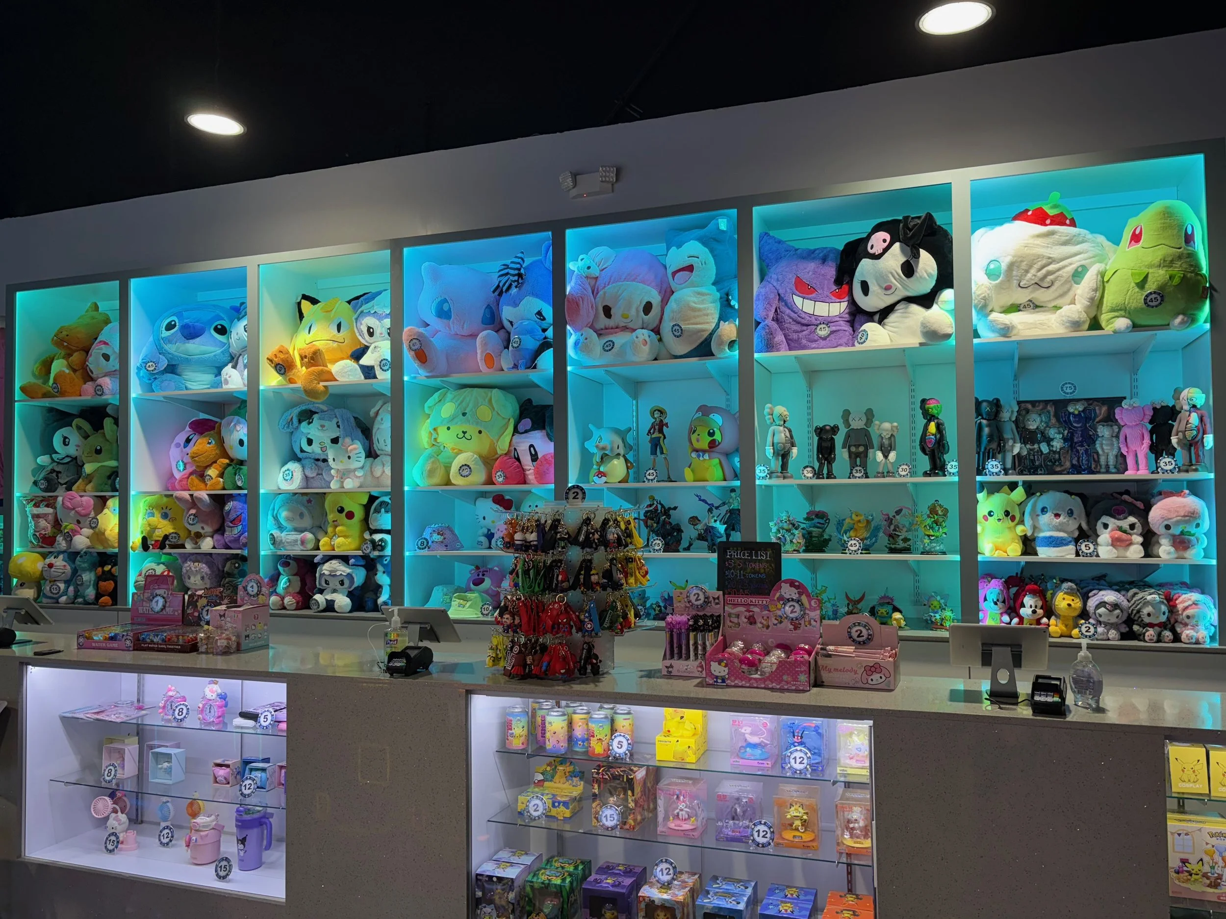 A toy store counter displaying various plush toys, collectibles, and keychains illuminated by colorful backlighting on shelves behind the counter.