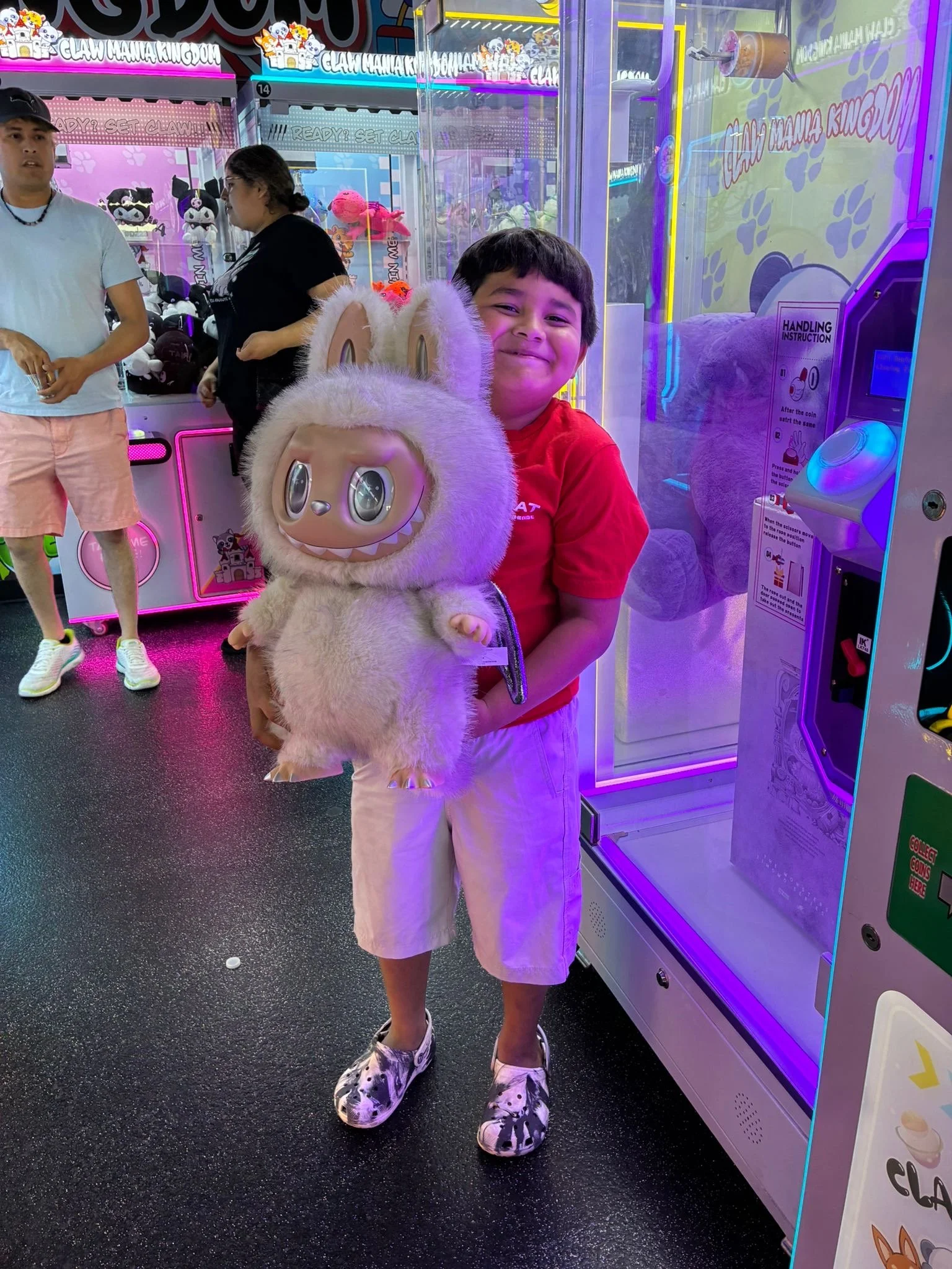 A boy holding a furry plush toy with large eyes and bunny ears in an arcade surrounded by claw machines and colorful lighting.