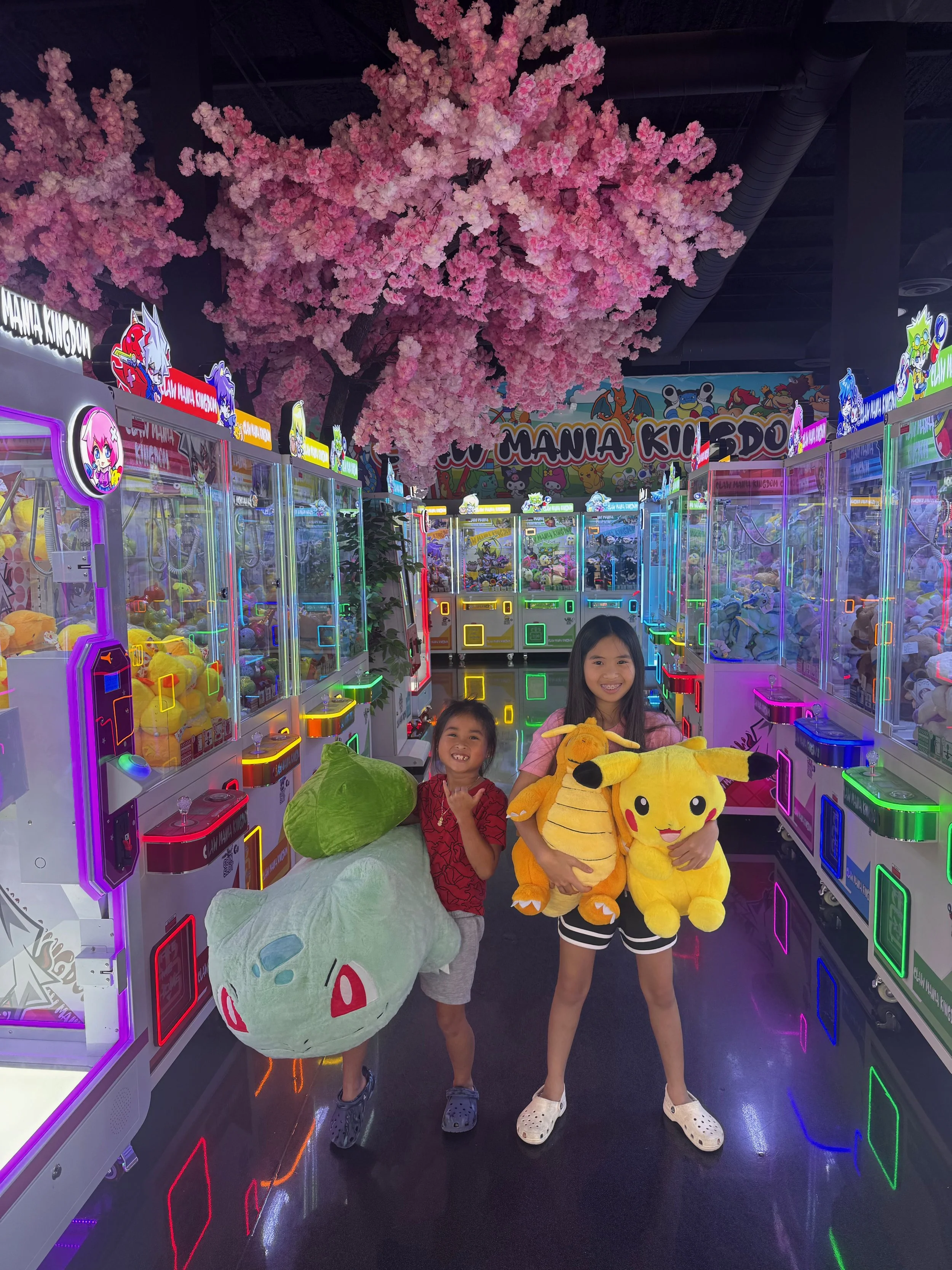 Two children holding large plush toys in an arcade filled with claw machines and colorful neon lights, with pink artificial cherry blossom trees overhead.