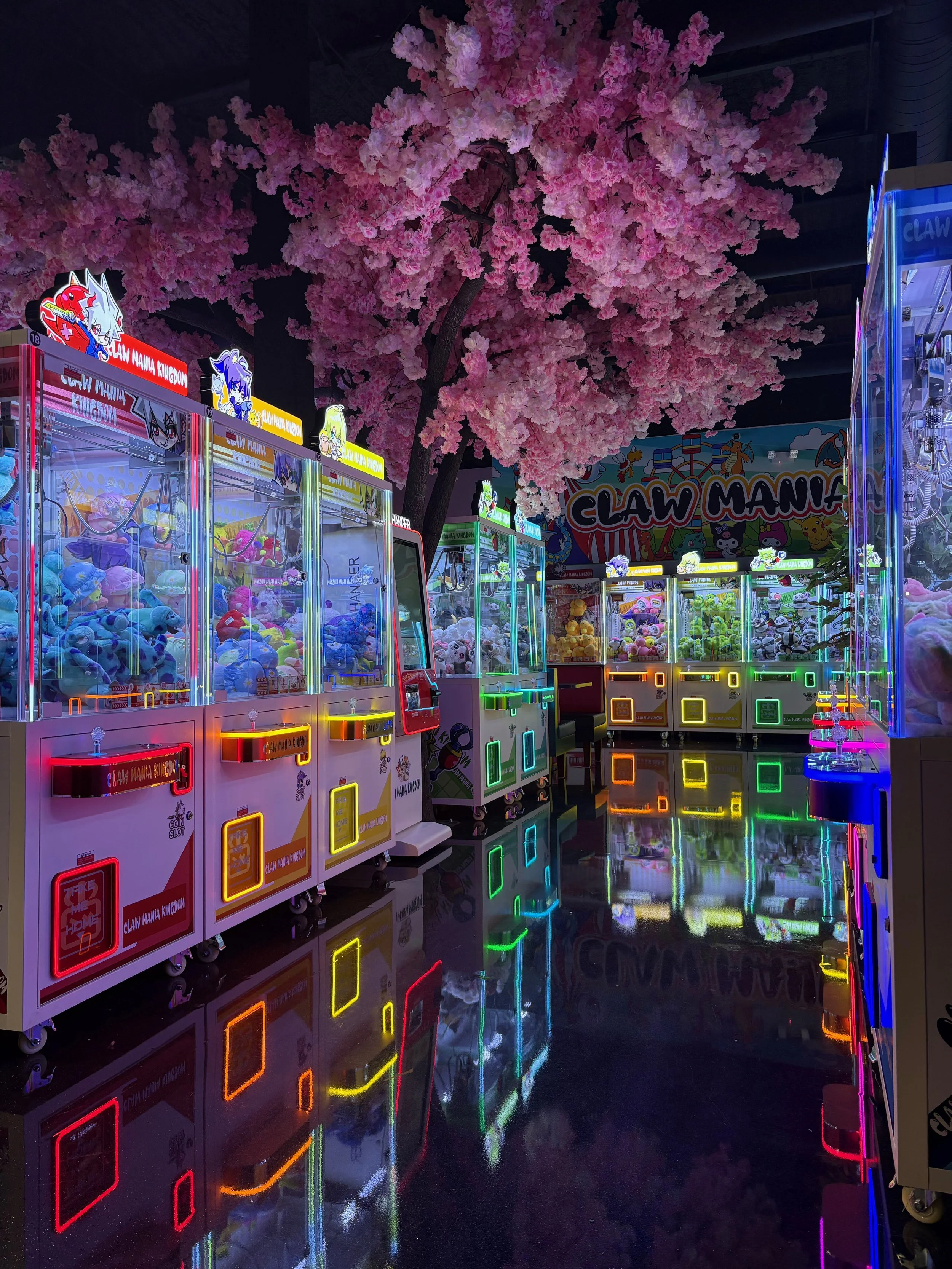 Colorful arcade with claw machines, neon lights, and a large pink cherry blossom tree, reflected on the shiny floor.