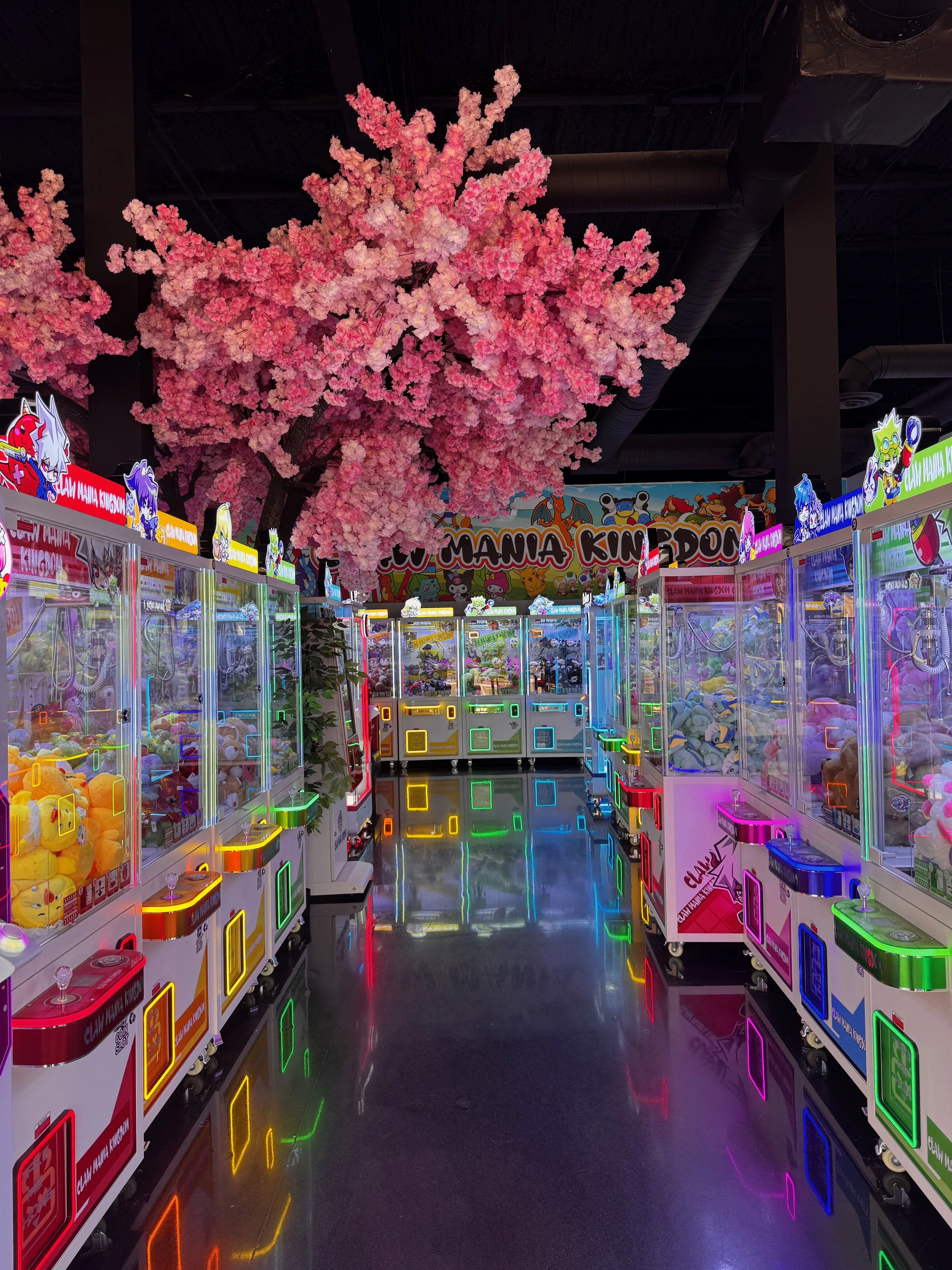 Indoor arcade filled with colorful claw machines, neon lights, and a decorative pink artificial cherry blossom tree at the center.