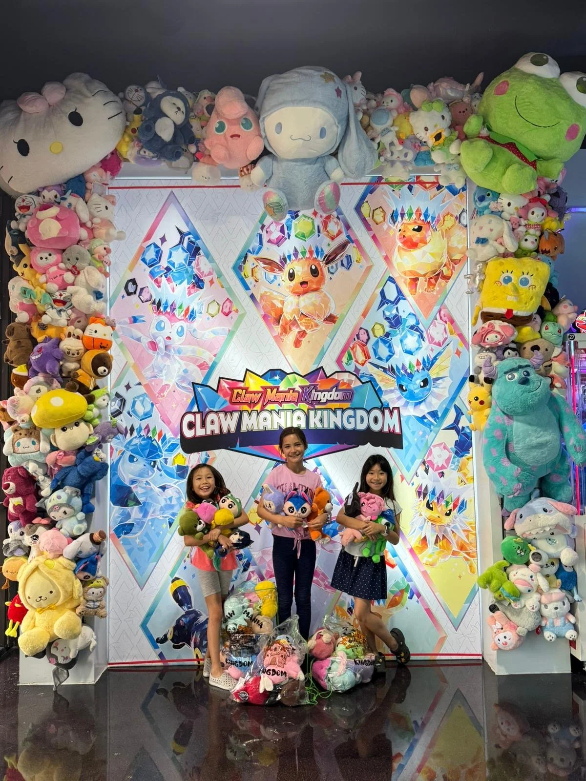 Three girls smiling and holding plush toys in front of a colorful background with the words 'Claw Mania Kingdom,' surrounded by numerous stuffed animal toys and decorations.