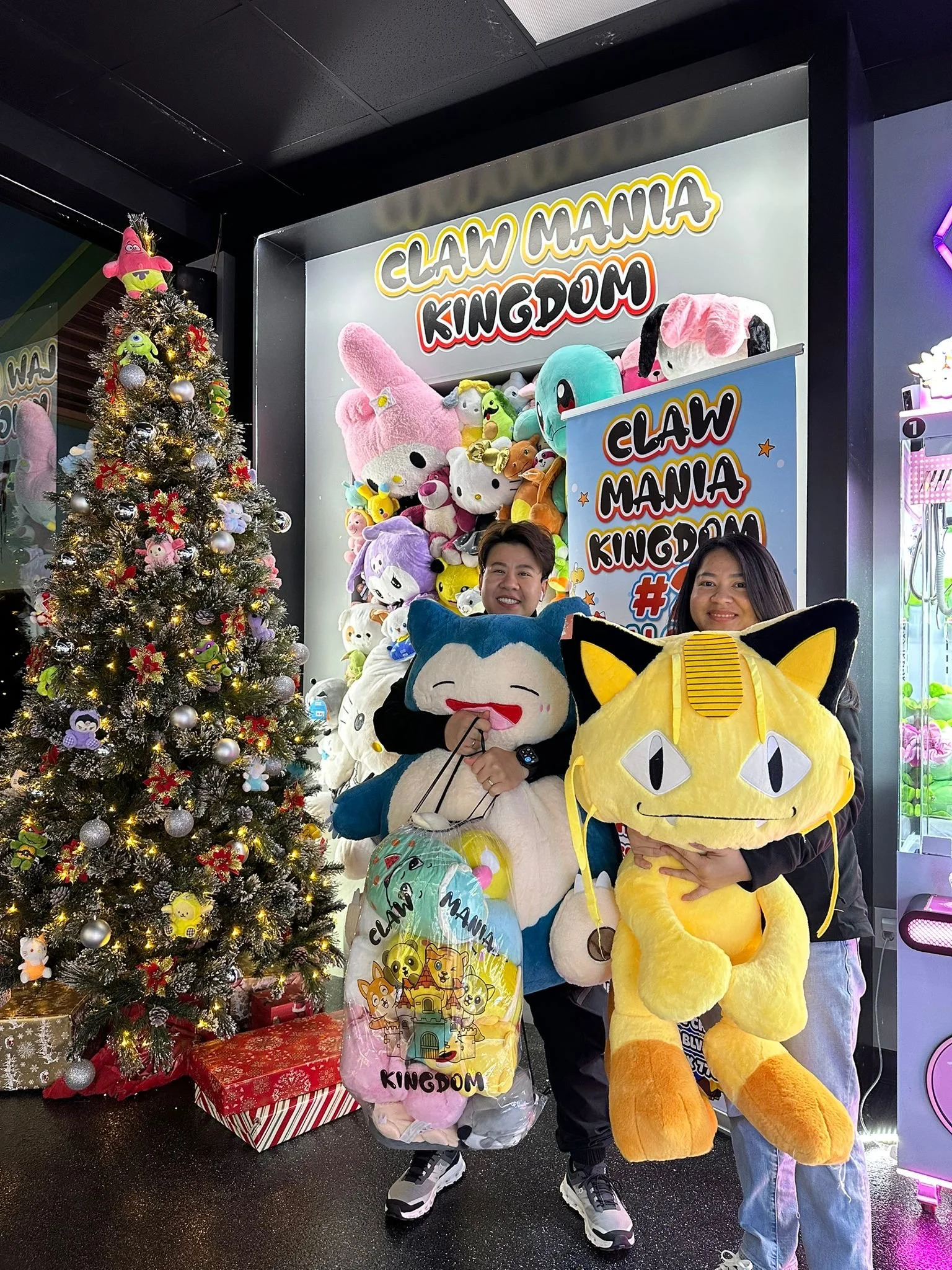 Two people holding large plush toys, standing beside a decorated Christmas tree with presents underneath, in front of a wall with a sign for Claw Mania Kingdom filled with plush toys.
