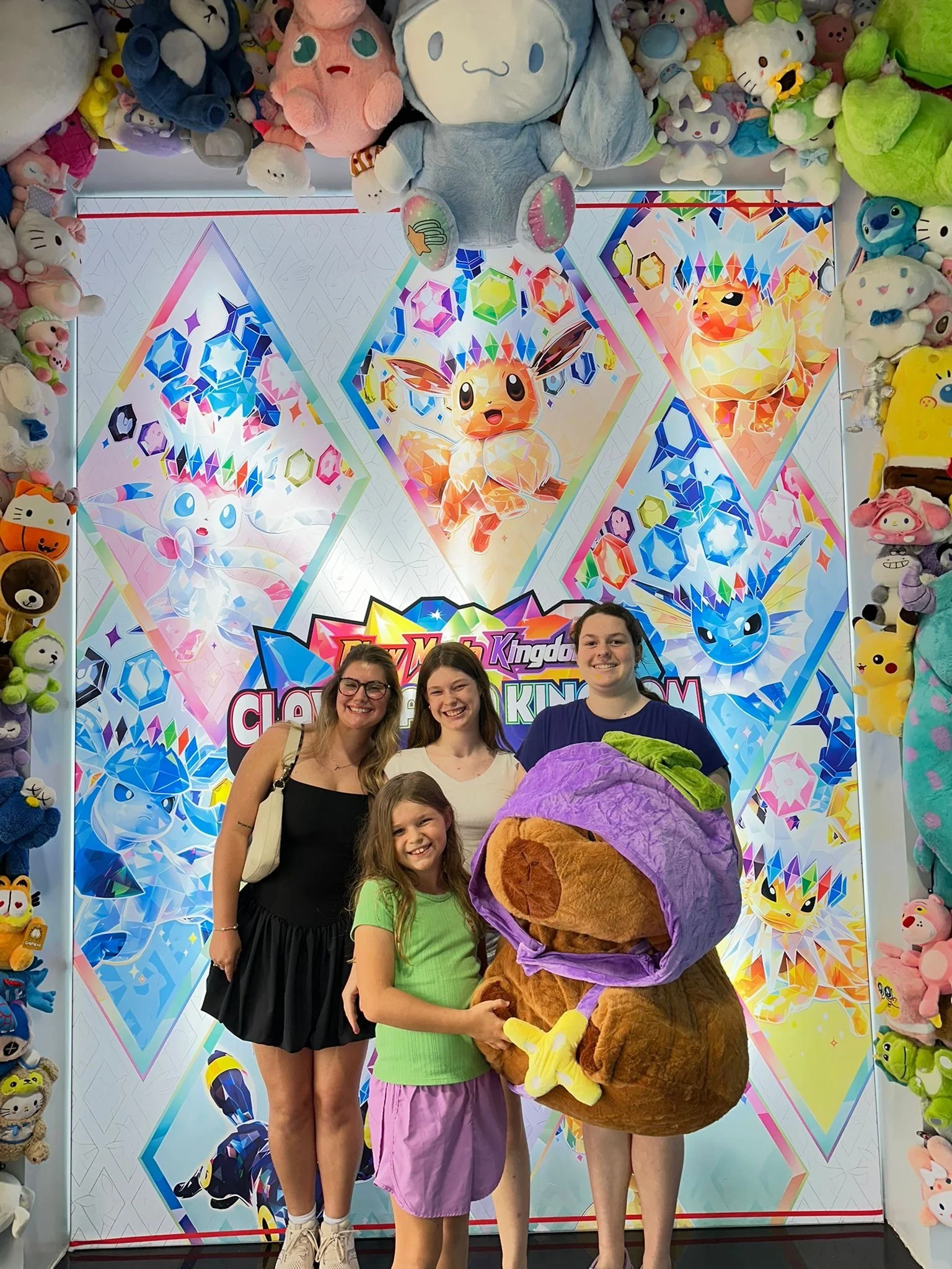 Four people, three young women and a girl, pose with a large plush toy of a brown Pokémon with a purple hat featuring a green leaf, in front of a colorful backdrop with Pokémon artwork and plush toys around the frame.