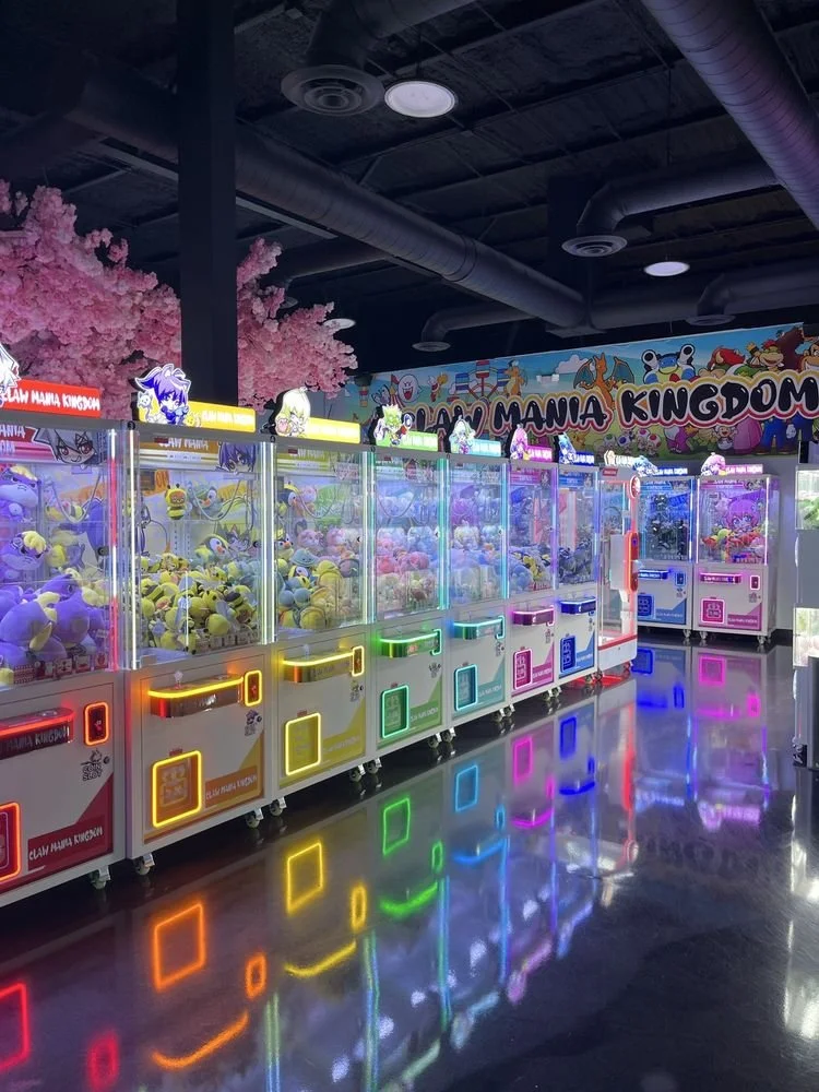Colorful claw machine arcade games filled with plush toys, with neon lights reflecting on the shiny floor, and pink ornamental trees in the background.