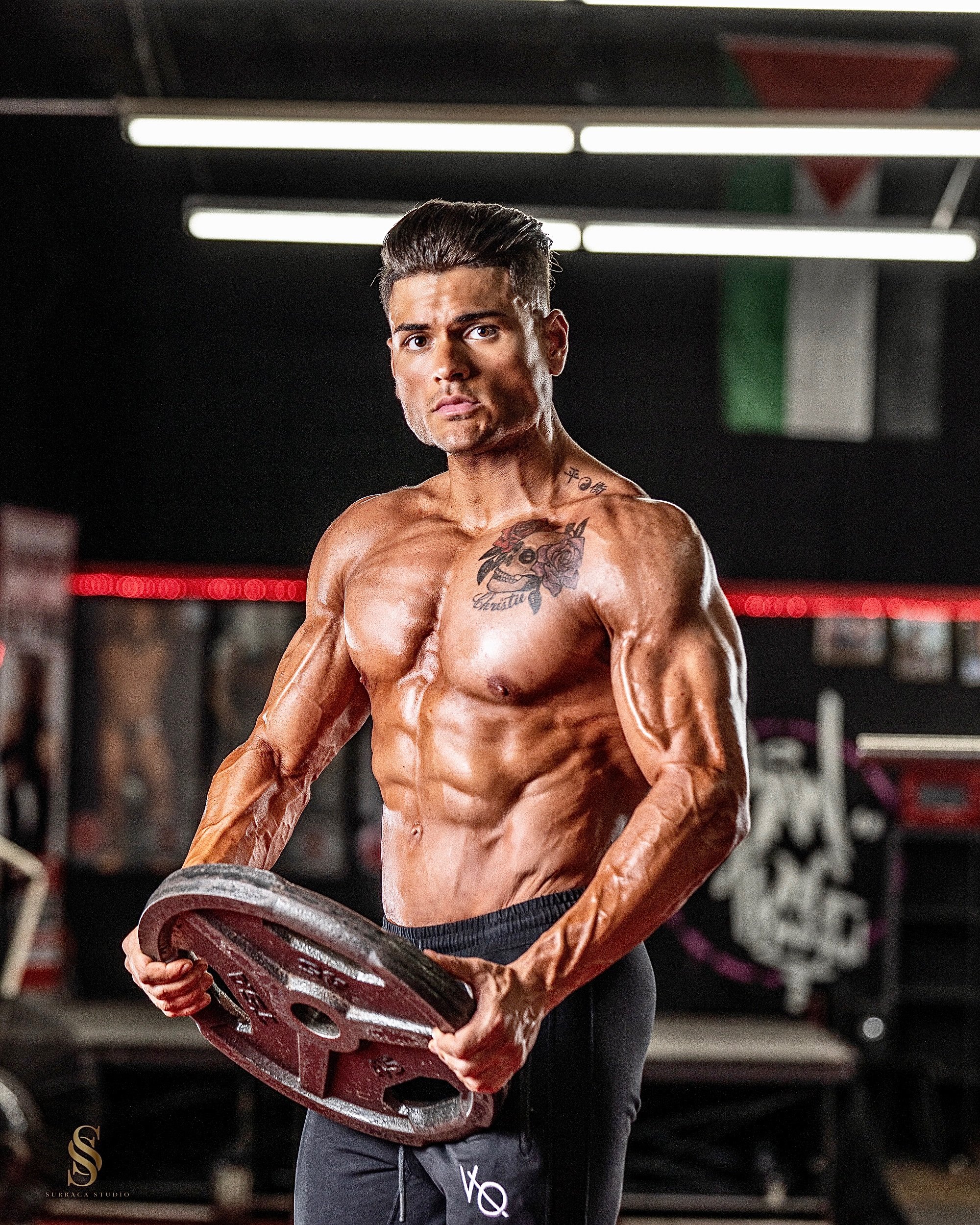 A shirtless man with muscular physique holding a weight plate in a gym, with tattoos on his chest and neck, and a serious expression.