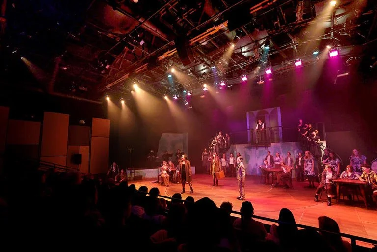 Theater stage with actors performing under colorful stage lights, with audience visible in the foreground.